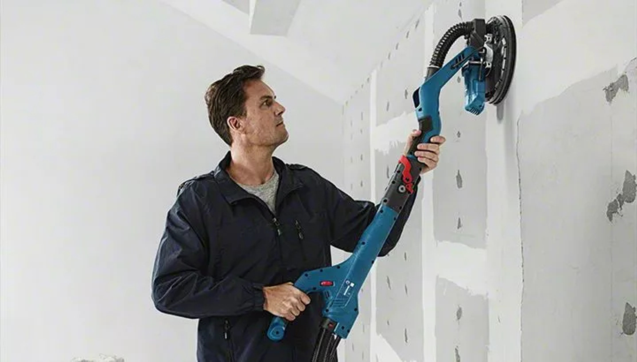 Homme ponçant des cloisons sèches sur un mur avec une ponceuse électrique bleue à long manche. Mur blanc, mur gris.