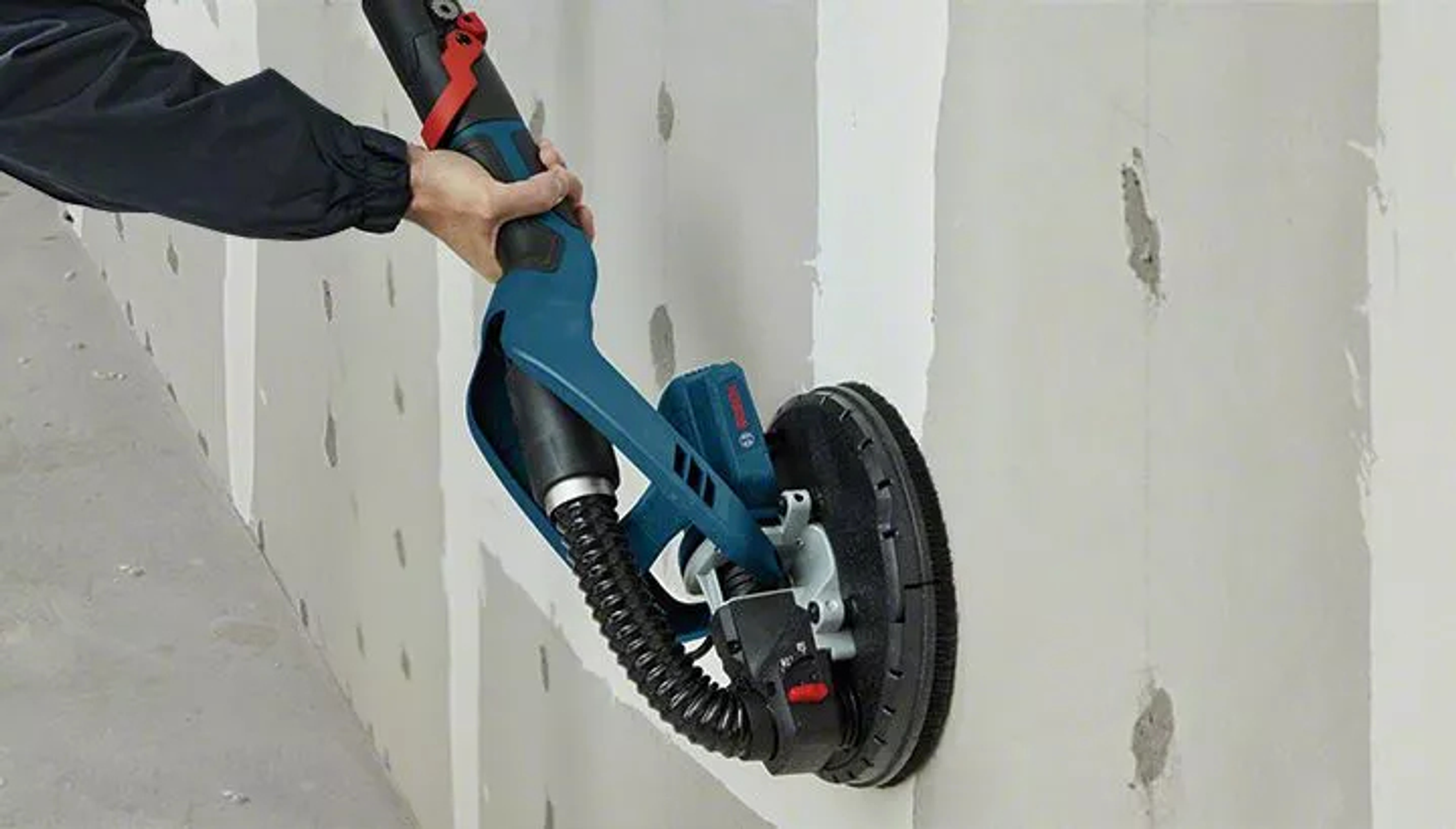 Une personne ponçant un mur avec une ponceuse à cloison sèche Bosch. La ponceuse est sur le mur, avec un tuyau.