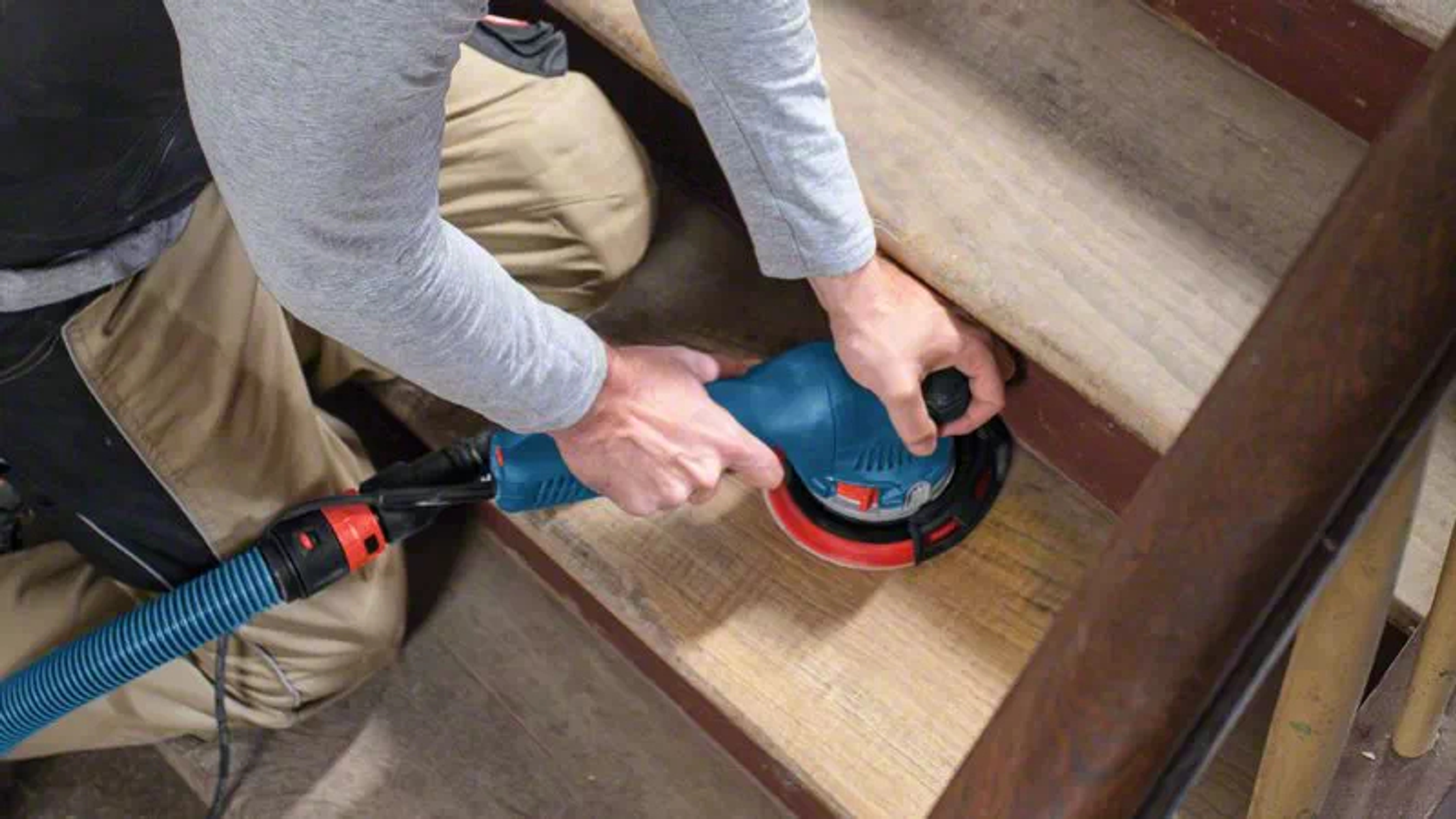 Una persona lijando escaleras de madera con una lijadora azul y negra, conectada a una manguera.