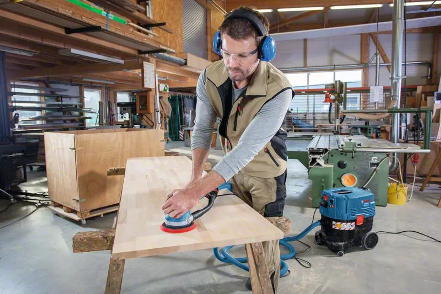 Hombre lijando una mesa de madera con una lijadora. Hay una aspiradora junto a él.