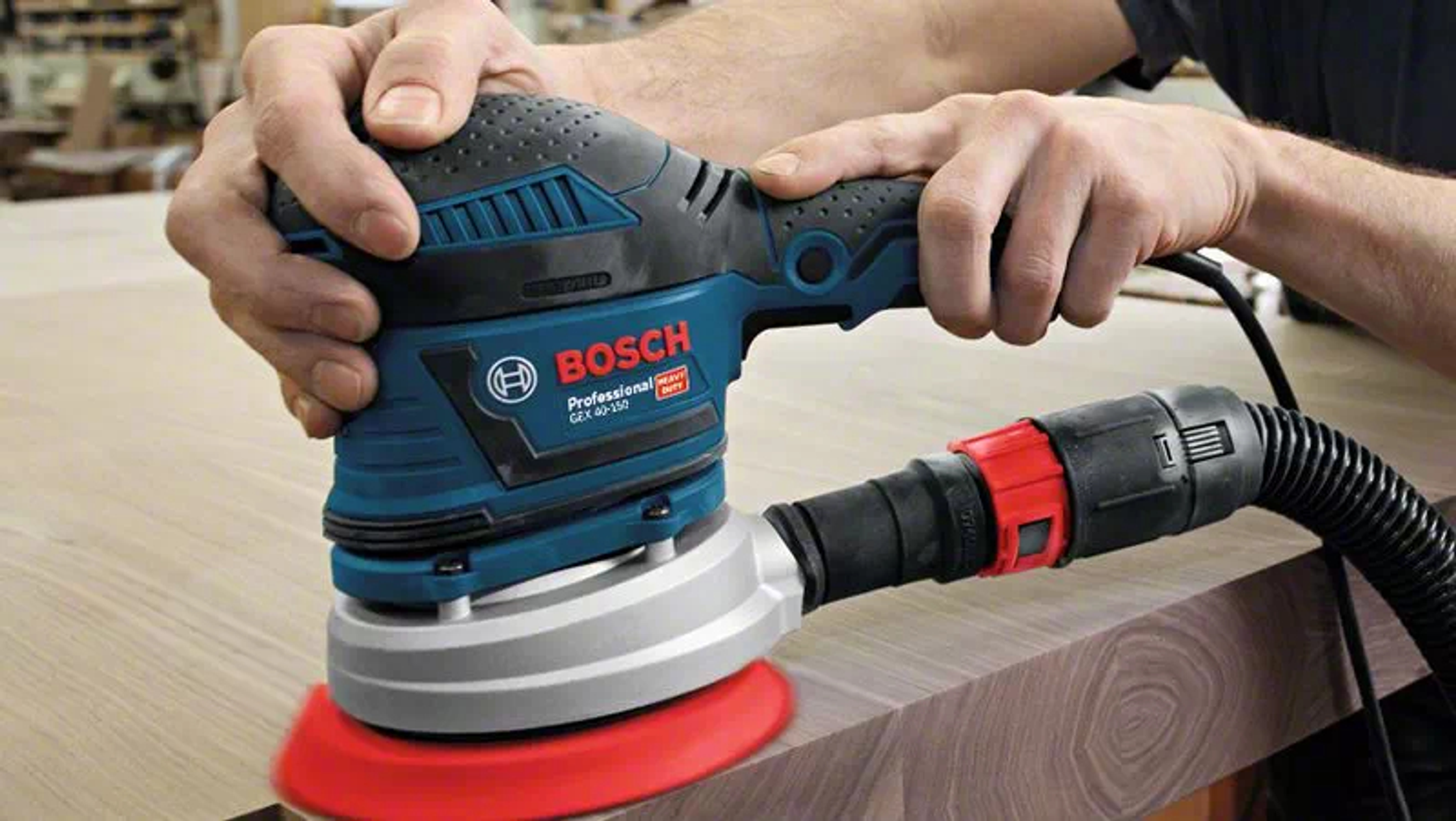 Hombre usando una lijadora Bosch en una superficie de madera. La lijadora es azul y negra.