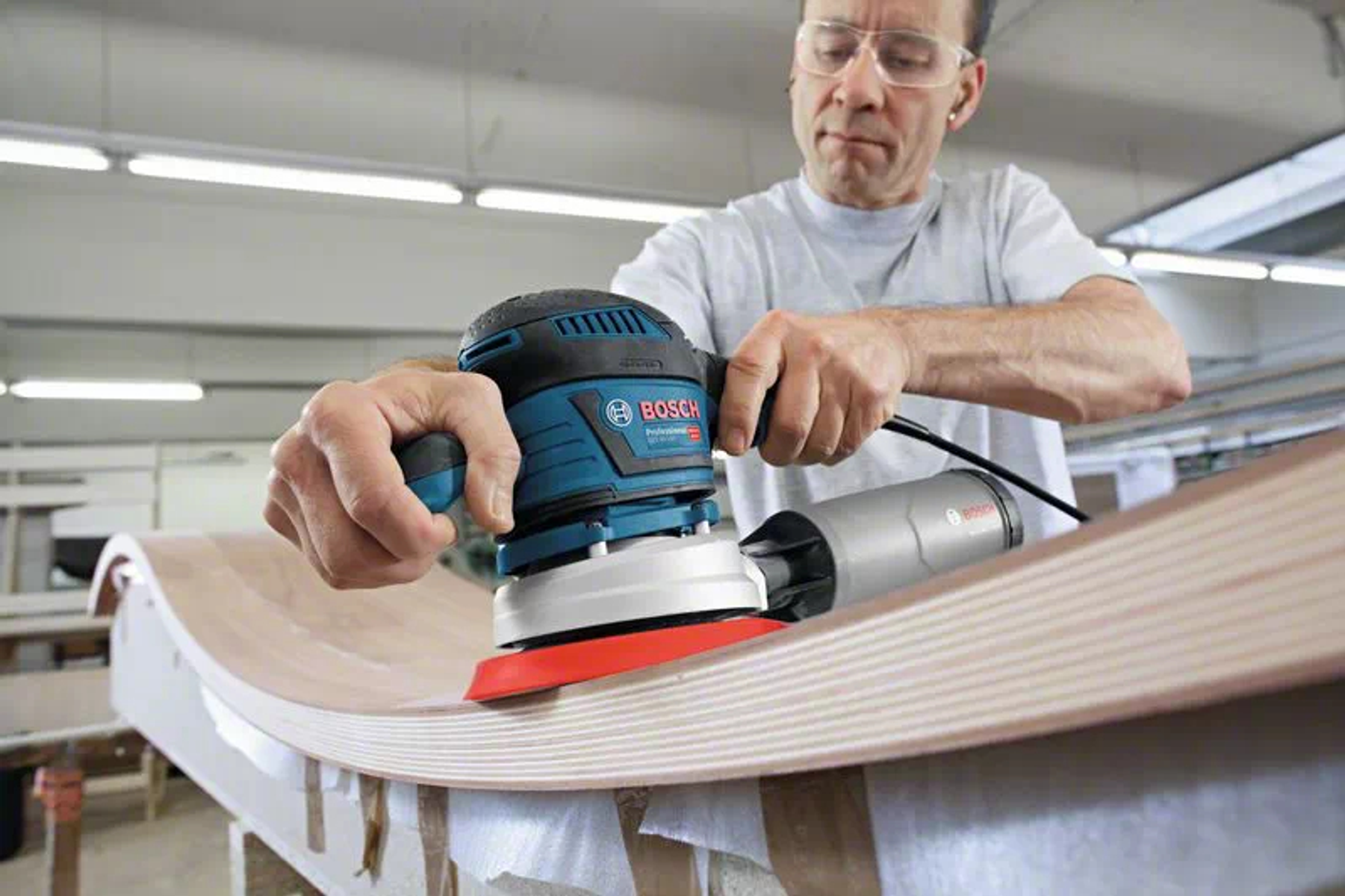 Hombre usando lijadora orbital azul y negra en superficie de madera curvada, con gafas de seguridad.