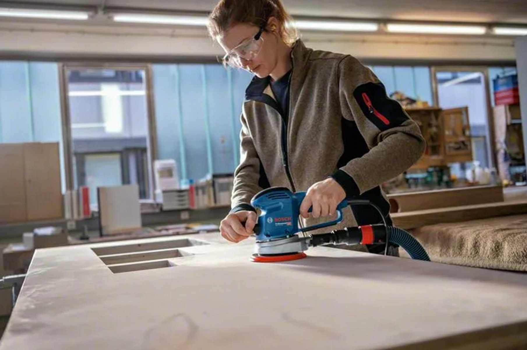 Una mujer lijando madera con una lijadora Bosch, usando gafas de seguridad, en un taller.