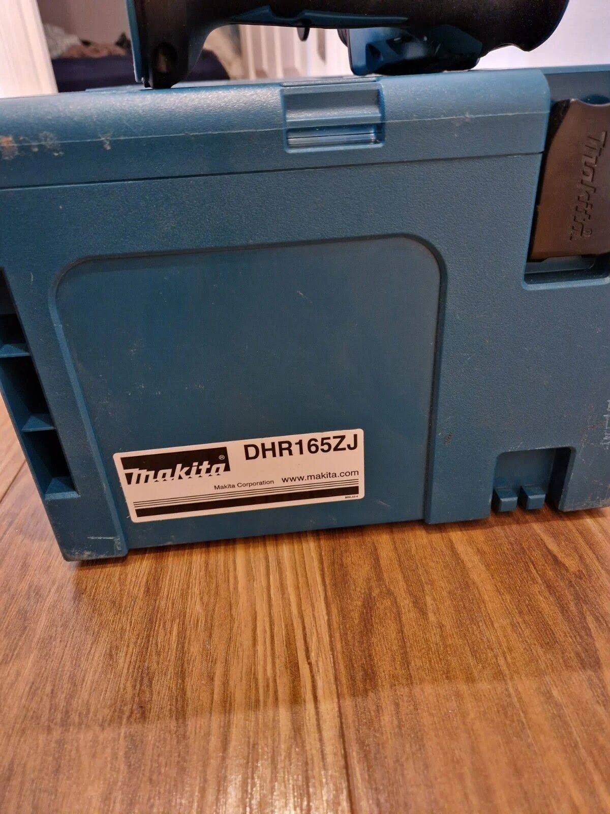 Una caja de herramientas Makita color turquesa con una etiqueta blanca que muestra 'DHR165ZJ' y el logotipo de Makita, sobre madera.