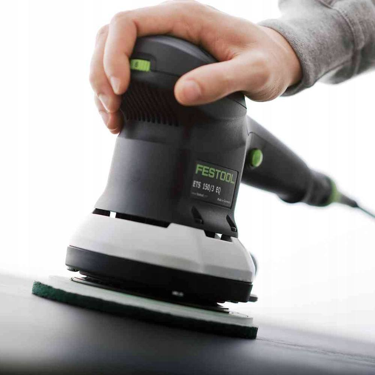 Mano sujetando una lijadora Festool, lijando una superficie oscura. La lijadora es negra y gris.