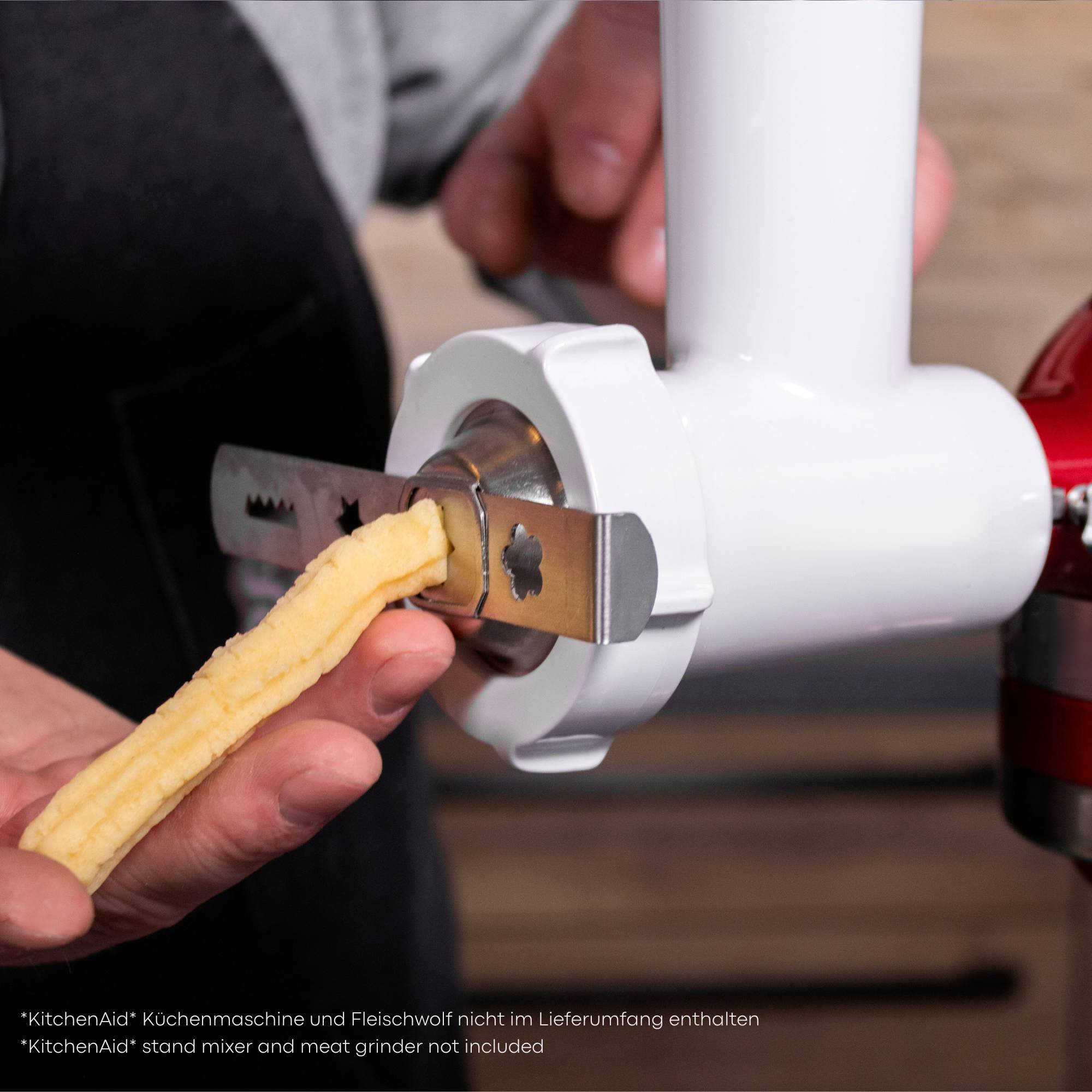 Hand hält gelbe Pasta, die in einen weißen KitchenAid-Fleischwolfaufsatz gefüttert wird, roter KitchenAid-Mixer im Hintergrund.