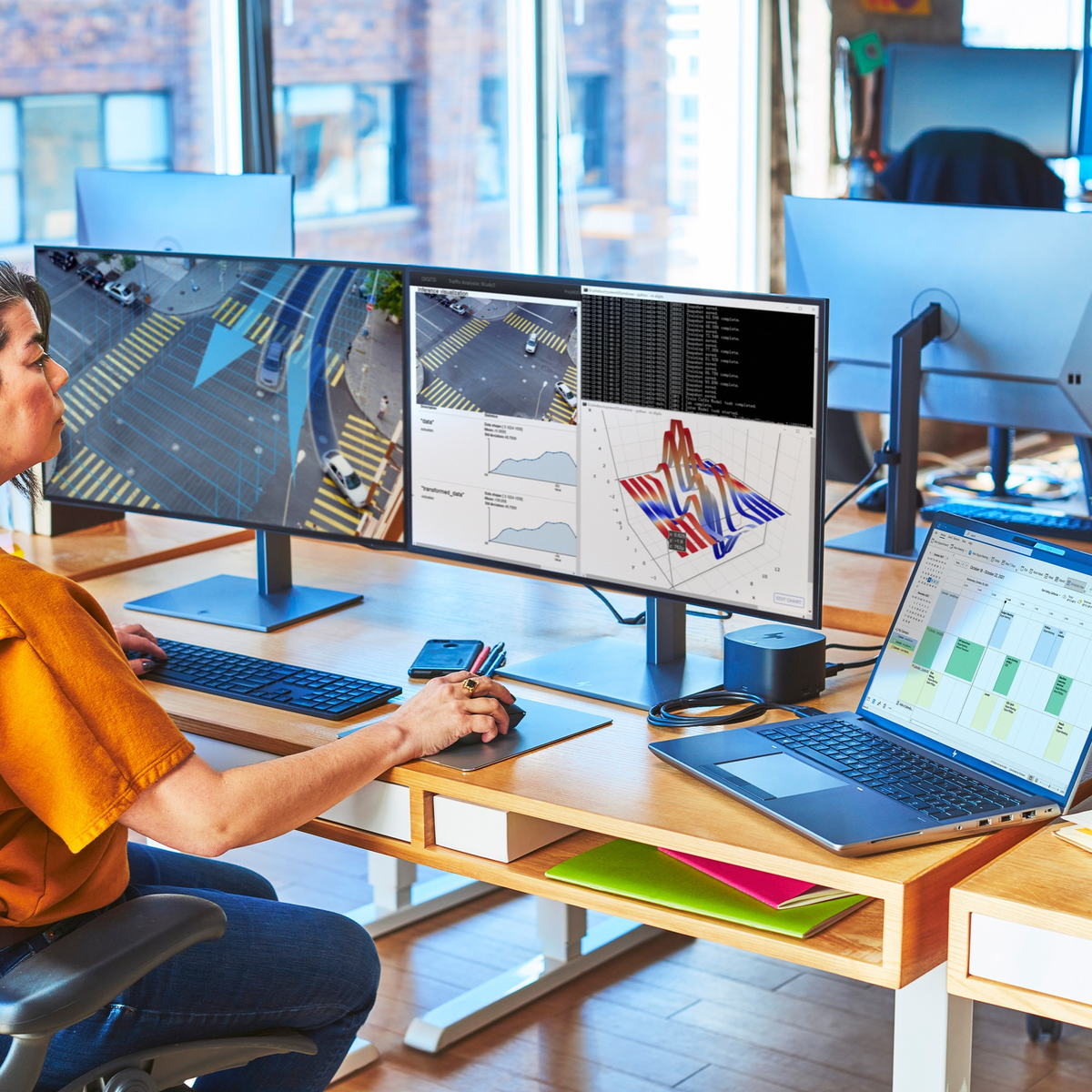 Osoba korzystająca z komputera z trzema ekranami i laptopem. Ekrany wyświetlają grafikę i dane.