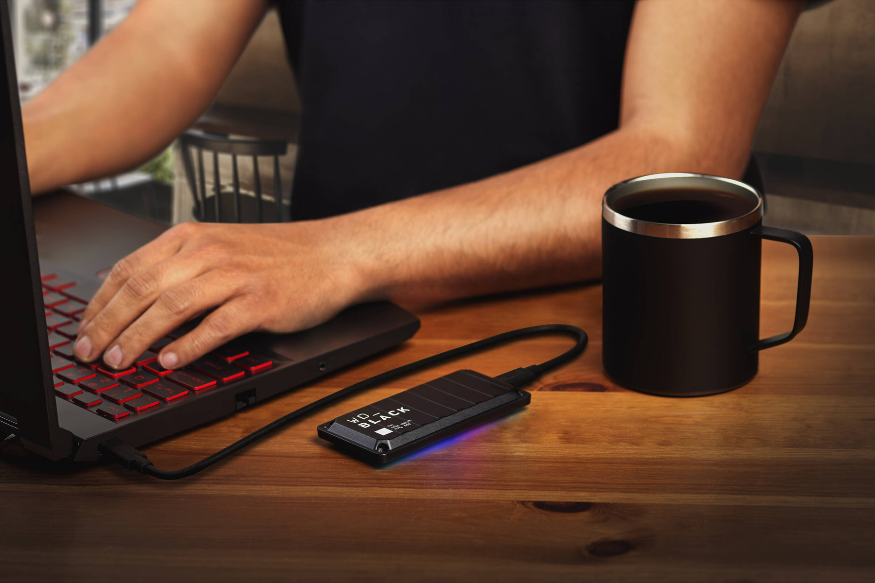 Osoba używa laptopa, podłączony dysk zewnętrzny. Kubek kawy na drewnianym stole.