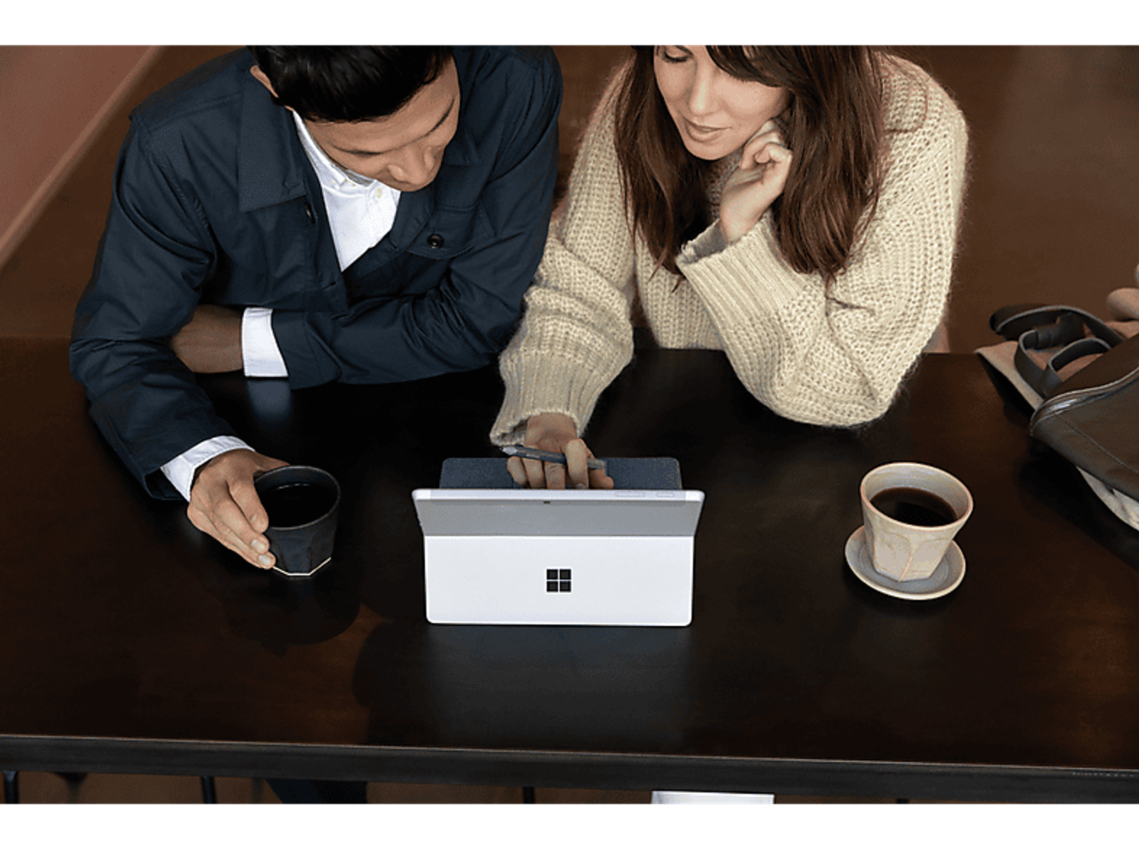 Zwei Personen an einem Tisch mit einem weißen Laptop und Kaffee. Sie schauen auf den Bildschirm.