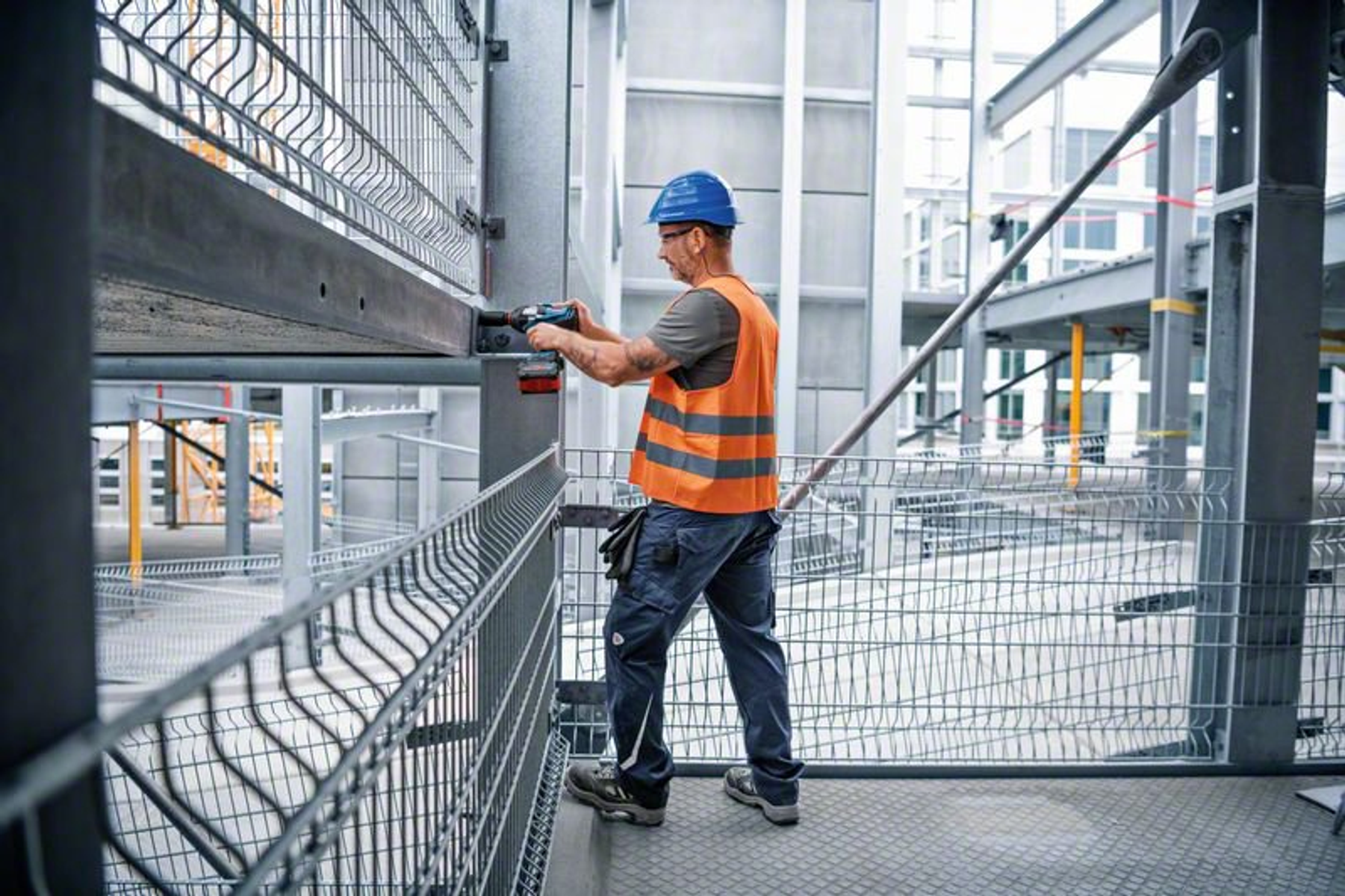 Robotnik budowlany w kamizelce i kasku ochronnym pracuje przy metalowej konstrukcji, używając elektronarzędzia.