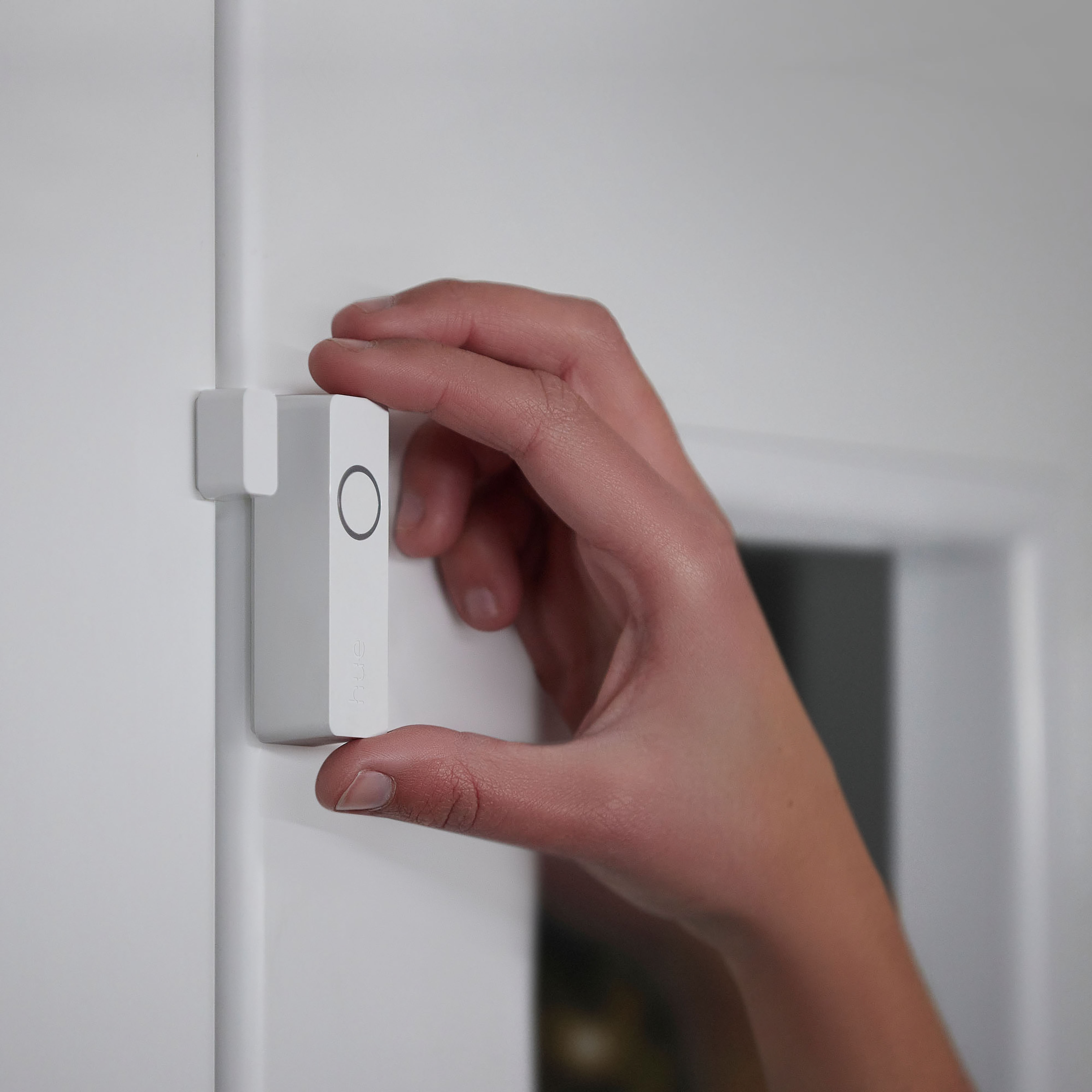 Ręka trzymająca biały czujnik inteligentnego domu. Czujnik znajduje się na białej ramie drzwi.