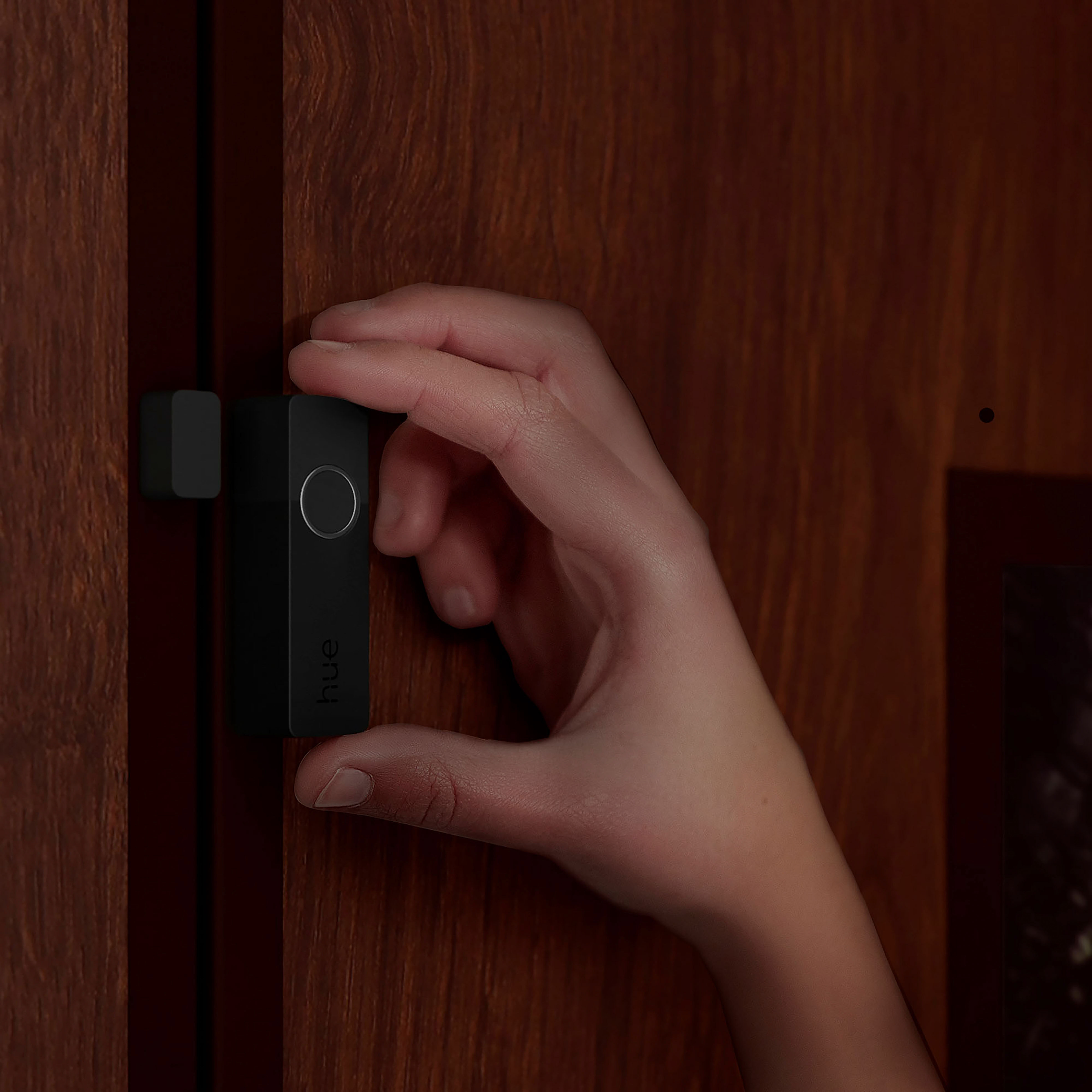 Ręka trzyma czarny czujnik przy drewnianych drzwiach. Na czujniku napis 'hue'.