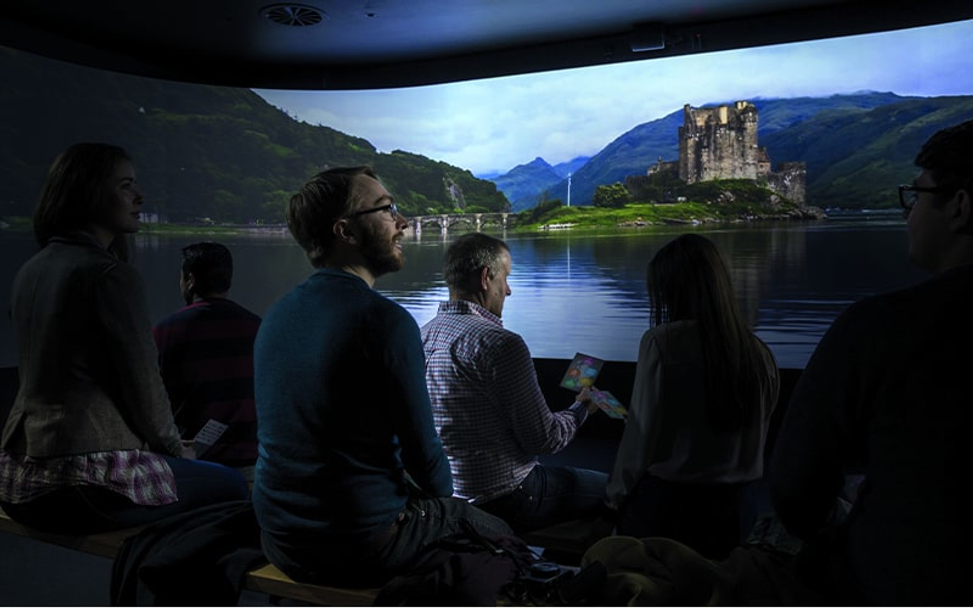 Personengruppe betrachtet projiziertes Bild eines Schlosses auf einem See. Dunkler Raum, großer Bildschirm.