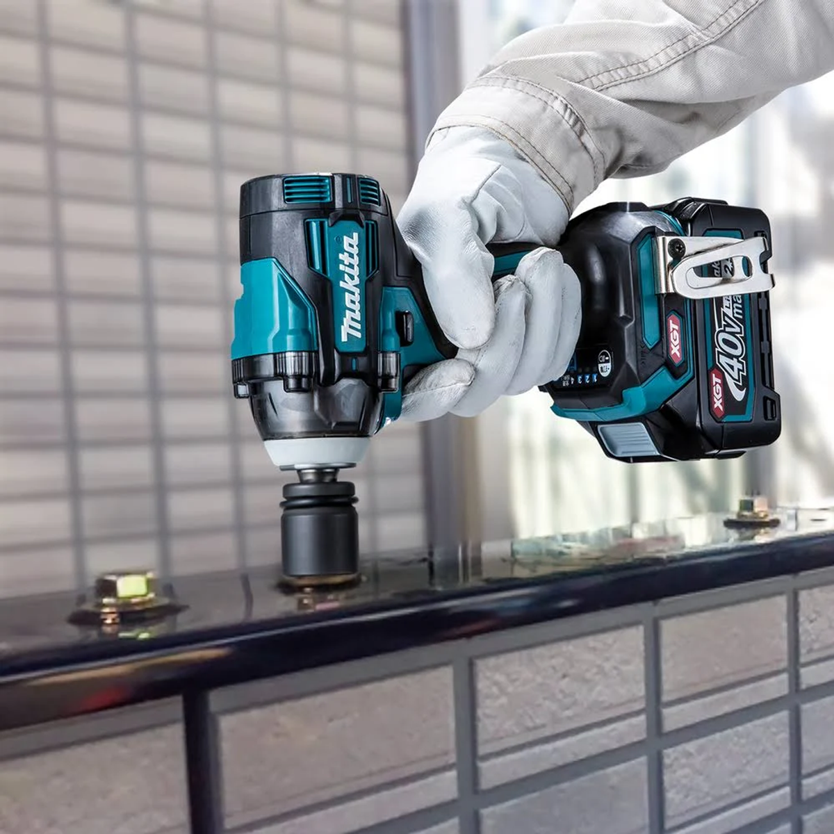 Un trabajador con guantes sostiene una llave de impacto Makita cerca de un riel metálico con pernos.