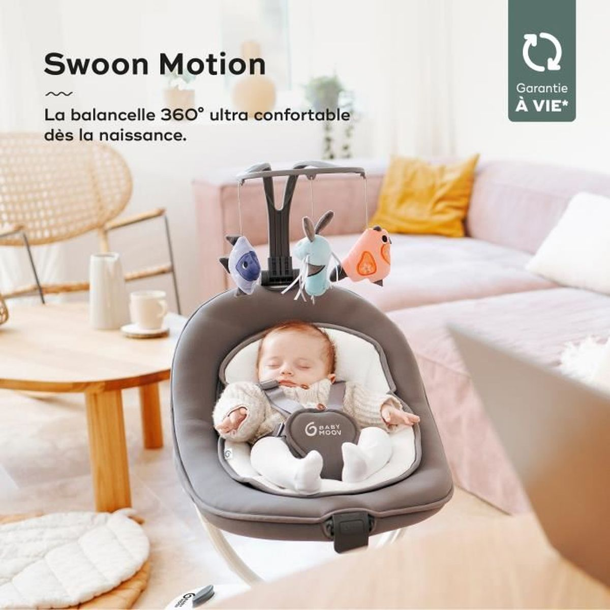 Baby in einer grauen Babyschaukel mit Spielzeug. Die Schaukel befindet sich in einem Haus mit Sofa und Holztisch.