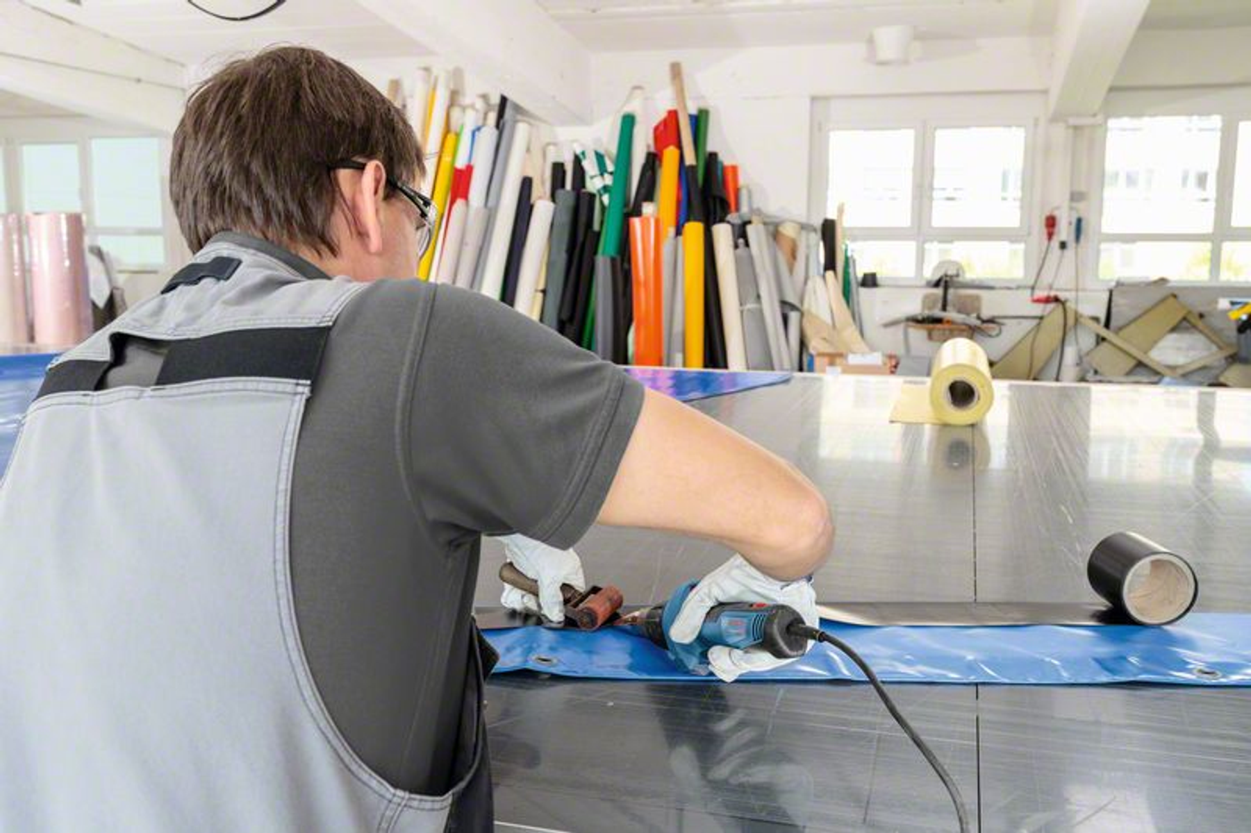 Osoba pracująca na niebieskiej plandece, używająca elektronarzędzia. Tło ma materiały.