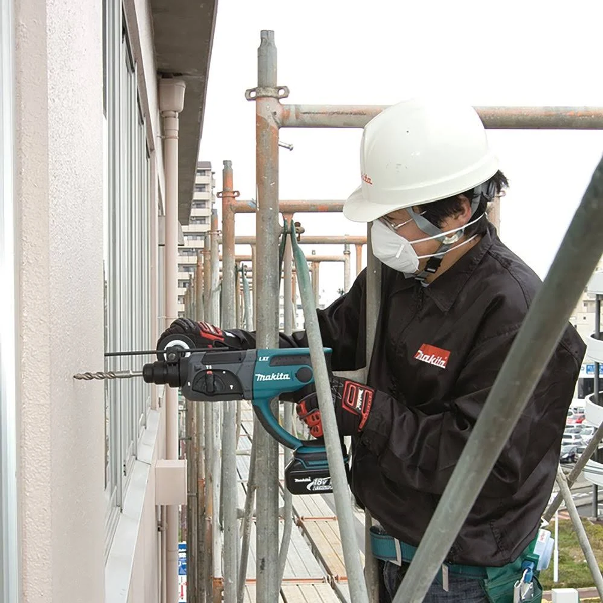 Trabajador taladrando una pared con un taladro Makita teal. Andamios y edificio al fondo.