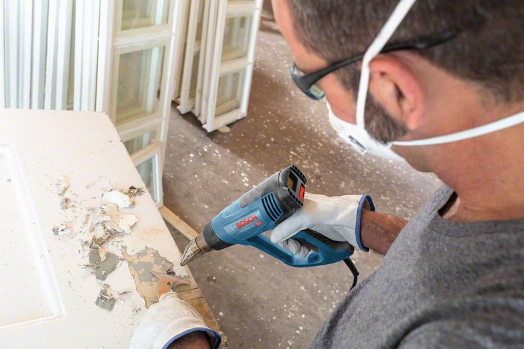 Mężczyzna w masce i rękawicach używa opalarki do usuwania farby z białych drzwi.