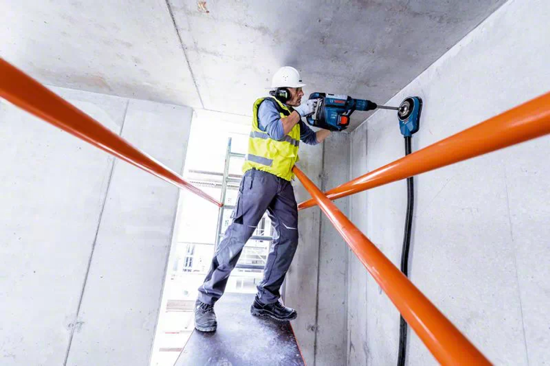Un trabajador de la construcción perforando una pared de hormigón. Está de pie en un andamio naranja.