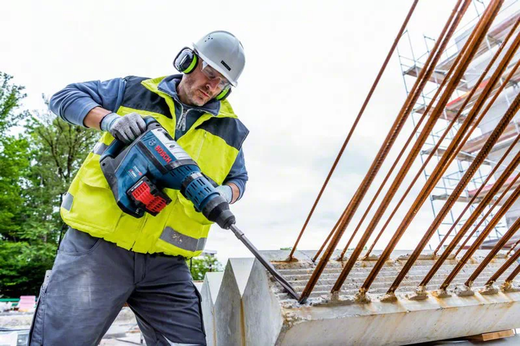 Un trabajador de la construcción usando un martillo demoledor Bosch. Usa chaleco amarillo, protección auditiva y gafas.