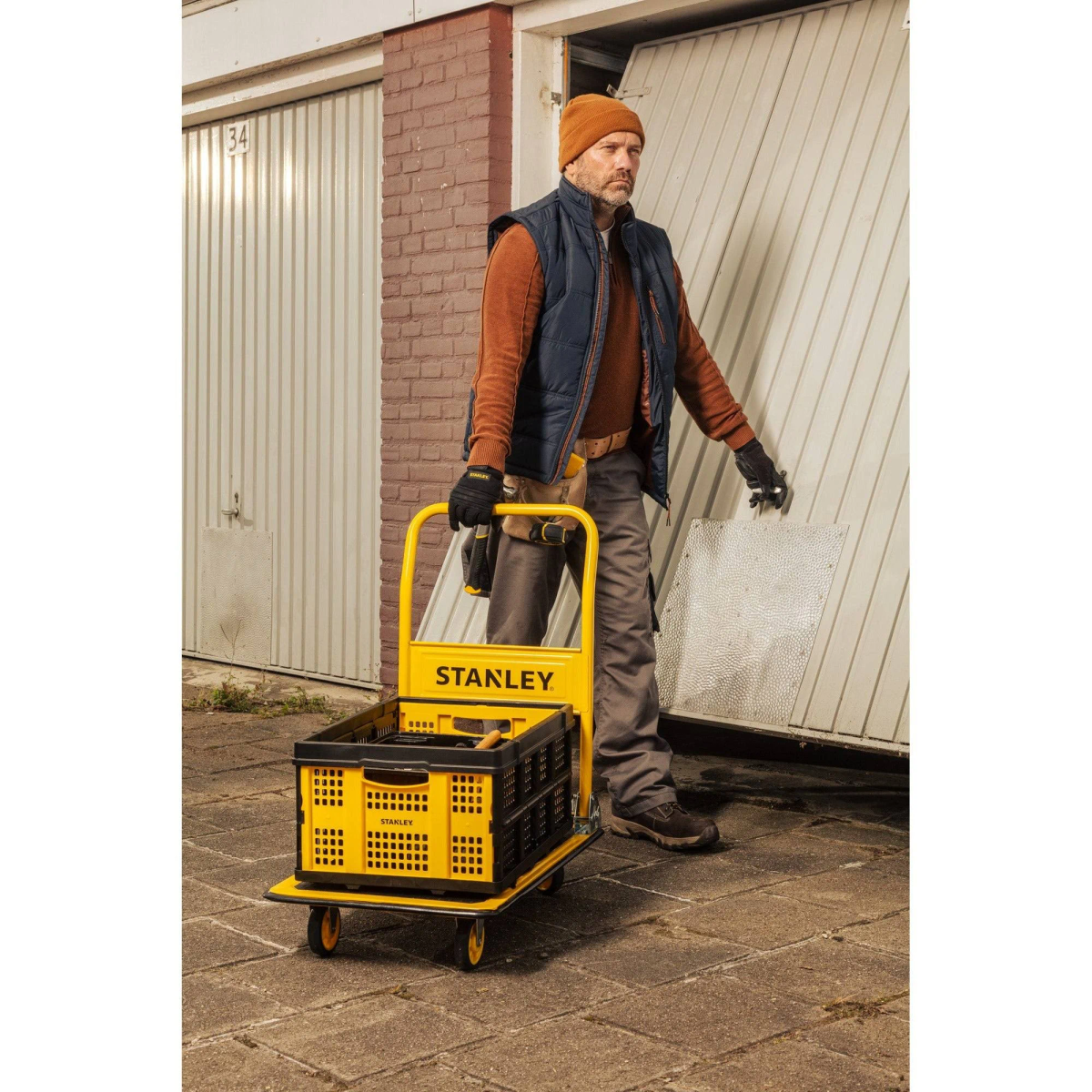 Man pushing a yellow Stanley cart carrying a black crate, near garage doors.