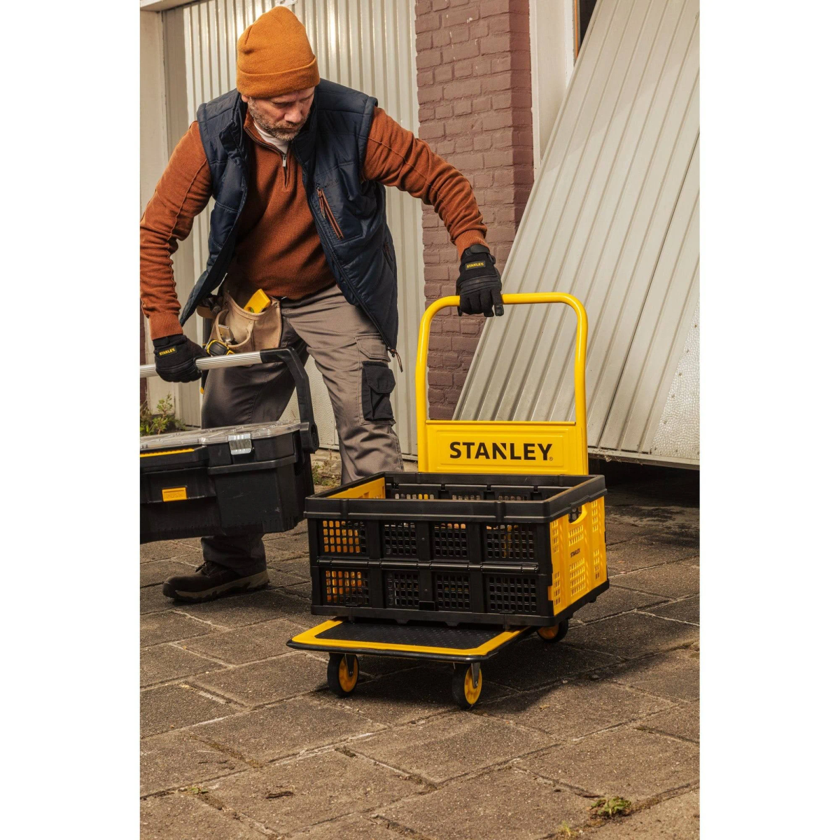 Man with a cart and black tool box. Cart is yellow and black. Brick wall in background.