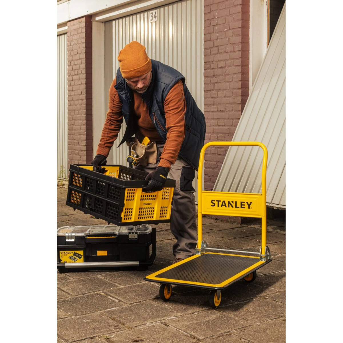 Man with a hat and a vest loading a black crate onto a yellow Stanley hand truck near a garage.