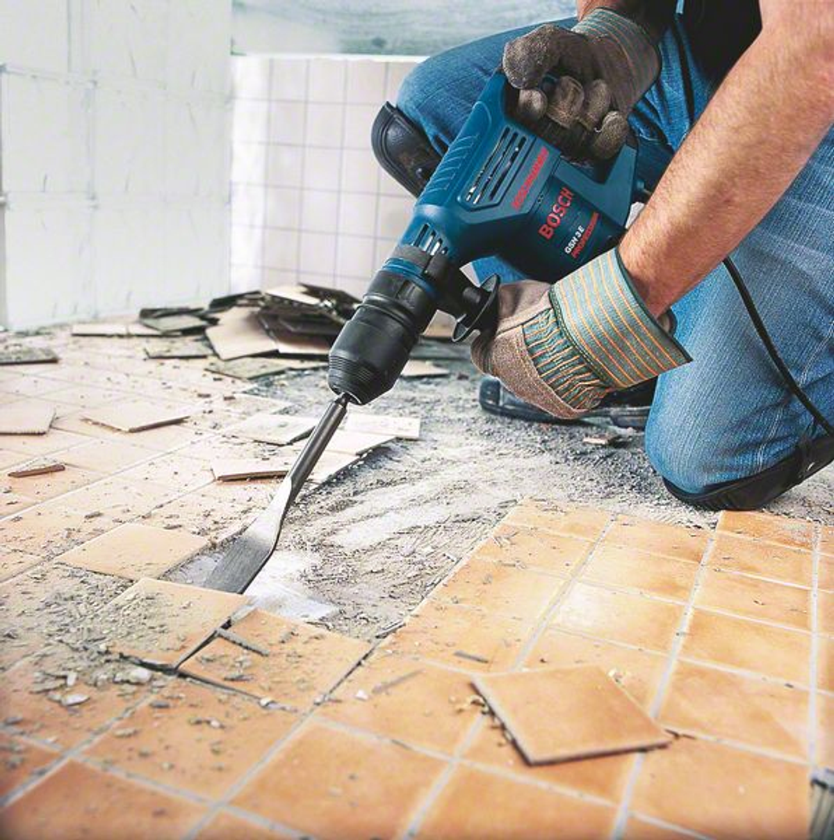 Hombre usando un martillo demoledor Bosch azul para romper baldosas en el suelo.