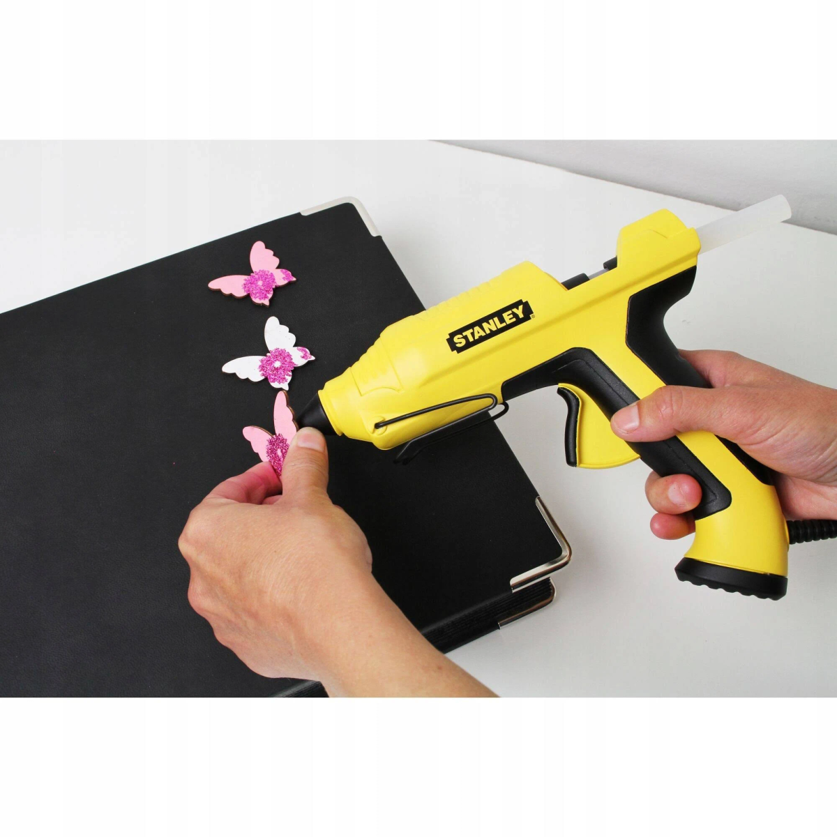 Persona usando una pistola de pegamento amarilla para pegar mariposas de purpurina rosa en una superficie negra.