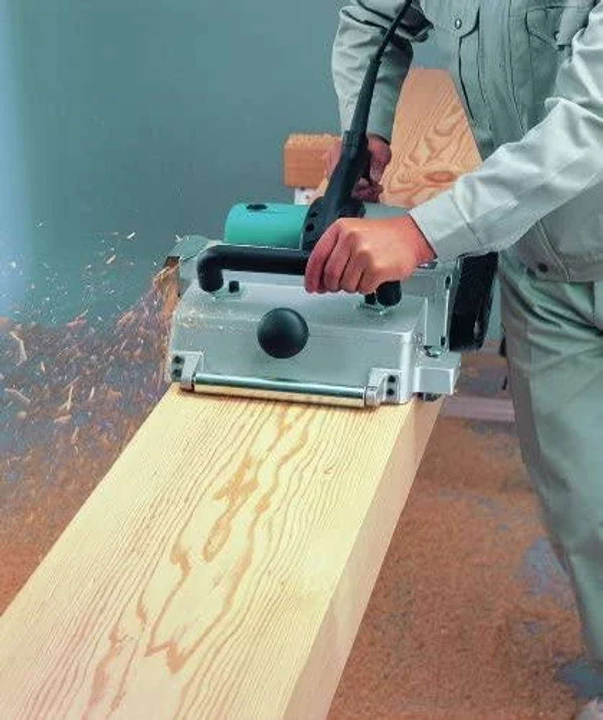 Un hombre usa una cepilladora plateada sobre una tabla de madera. Hay virutas en el aire. El fondo es turquesa.