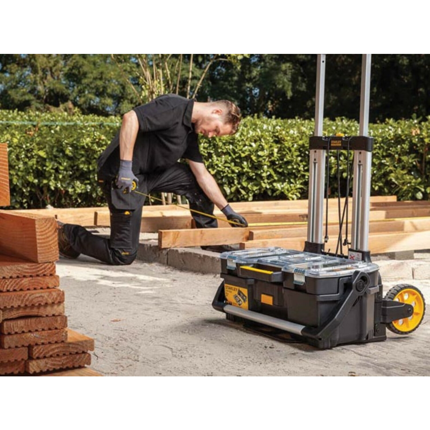 Man kneeling, measuring wood. Black and yellow rolling toolbox nearby. Outdoors.