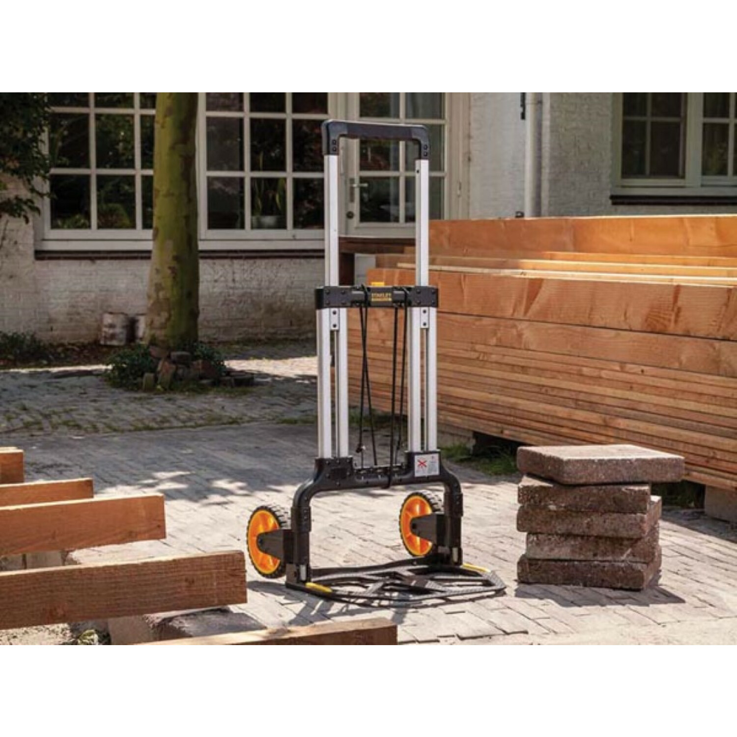 A hand truck with orange wheels stands near stacked bricks and a pile of lumber, with a building in the background.