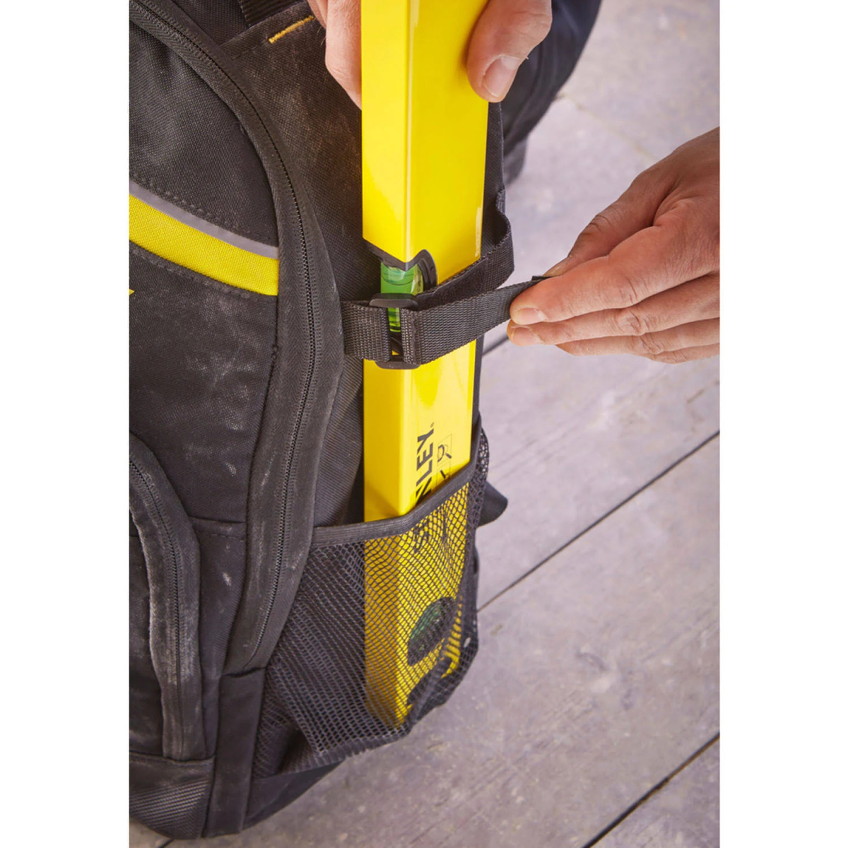 Manos de una persona asegurando un nivel amarillo en el bolsillo lateral de una mochila. Mochila negra y amarilla.