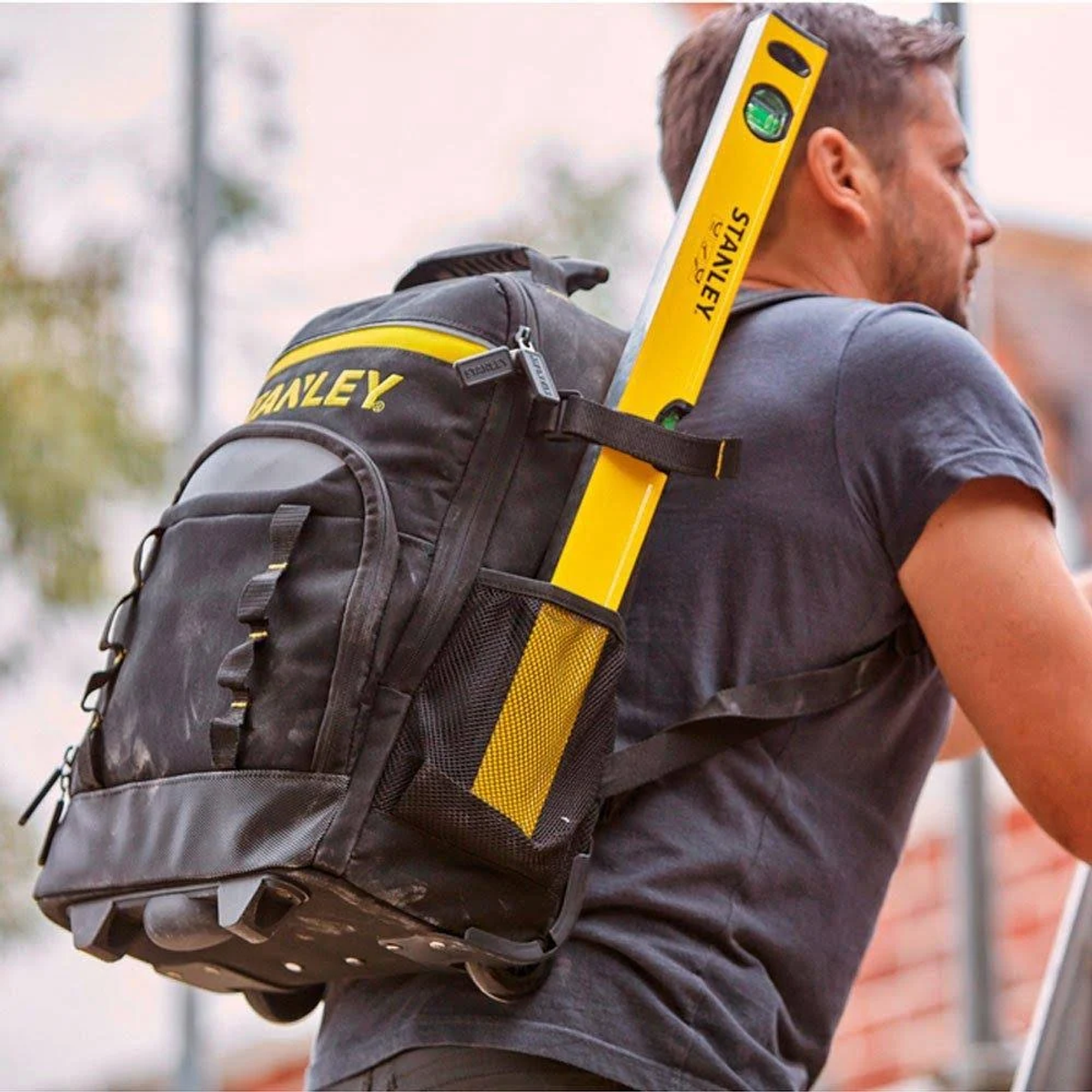 Hombre con mochila y nivel. Nivel amarillo adjunto. Mochila negra y amarilla. El hombre lleva una camisa gris.