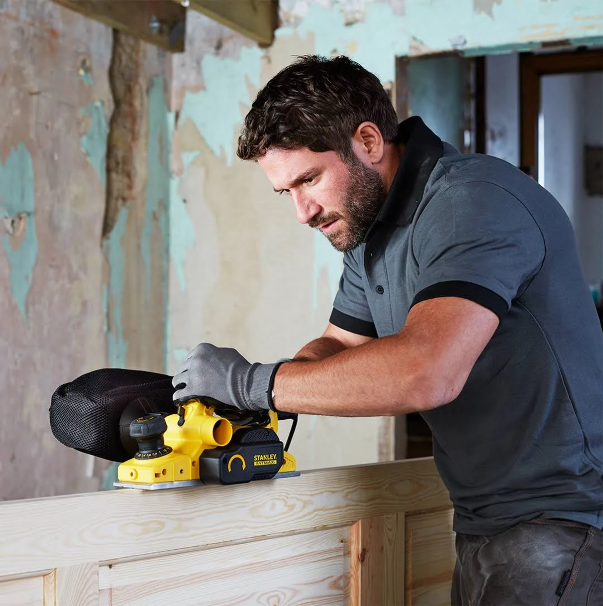 Hombre cepillando una puerta de madera. Usa una herramienta amarilla y negra. Interior.