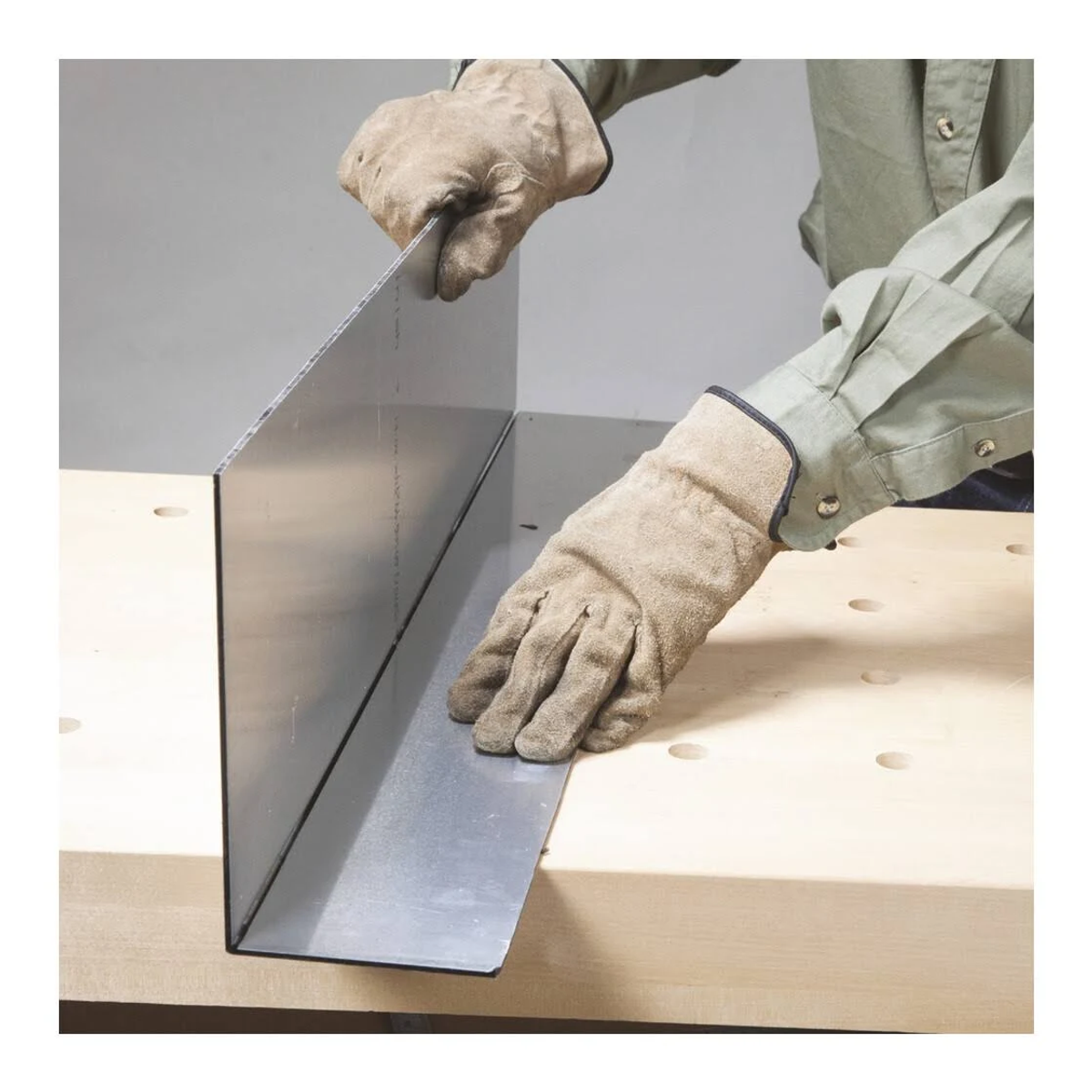 Mani che piegano un foglio di metallo su una superficie di legno. Sono visibili guanti.
