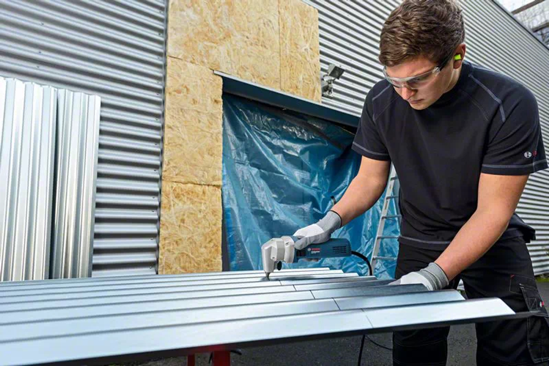 Un uomo con occhiali protettivi taglia una copertura metallica con un utensile Bosch, con protezione per le orecchie e guanti.