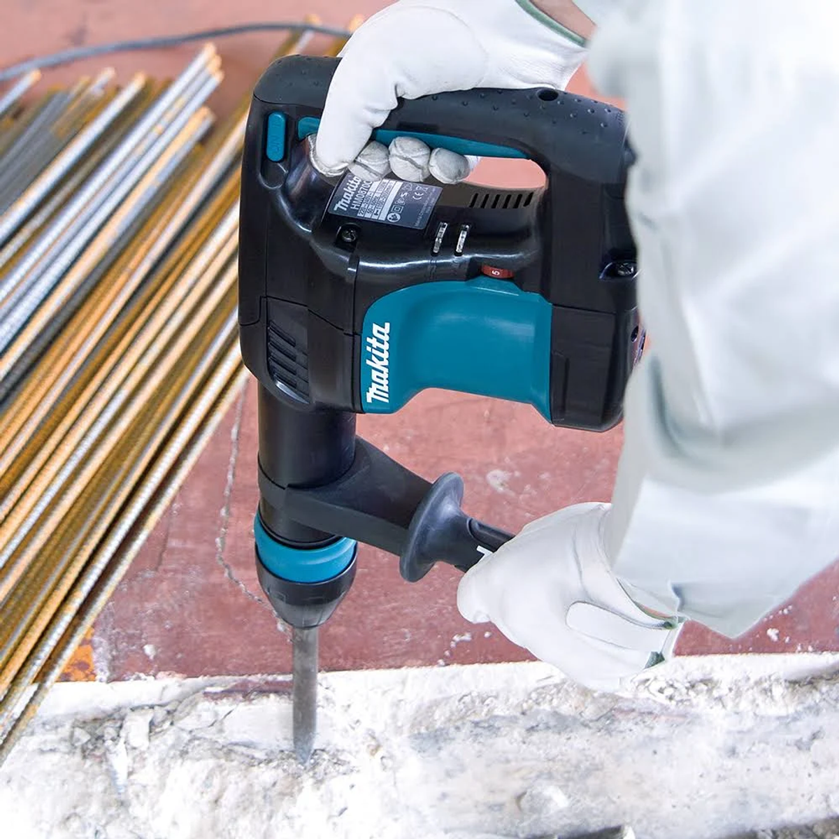 Una persona con guantes blancos usando un martillo perforador Makita para romper concreto, varilla de refuerzo en el fondo.