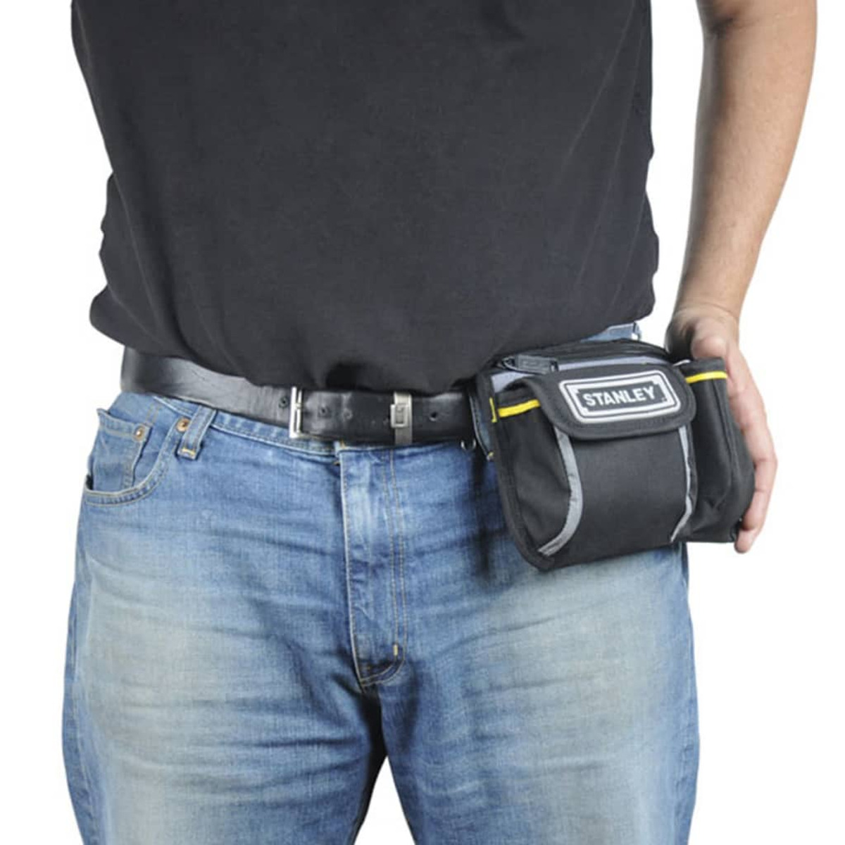 Un hombre con cinturón de herramientas negro con logo Stanley, de pie, jeans, fondo blanco.