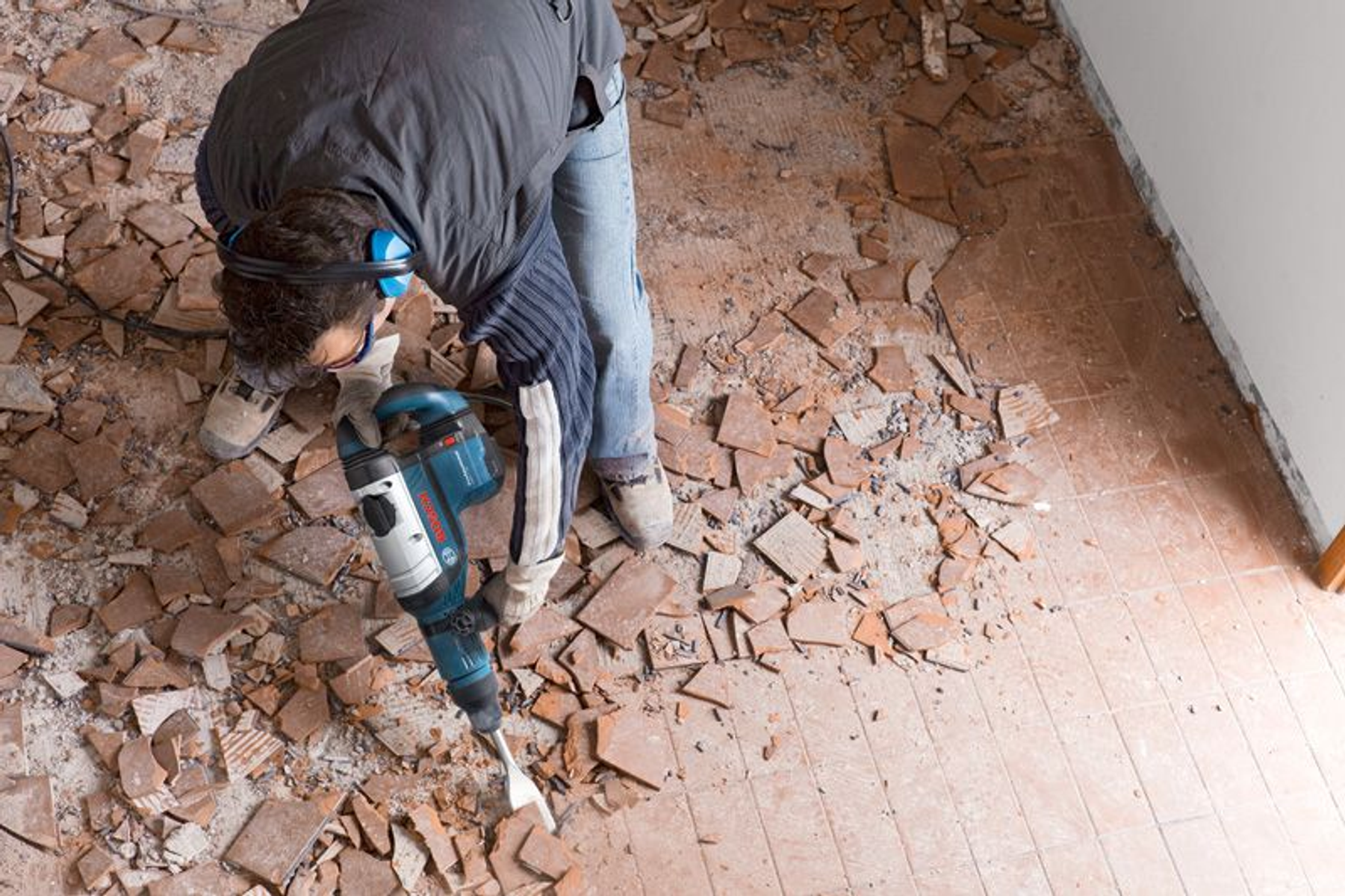 Una persona con equipaggiamento protettivo usa un martello demolitore Bosch su un pavimento piastrellato, circondato da piastrelle rotte e detriti.