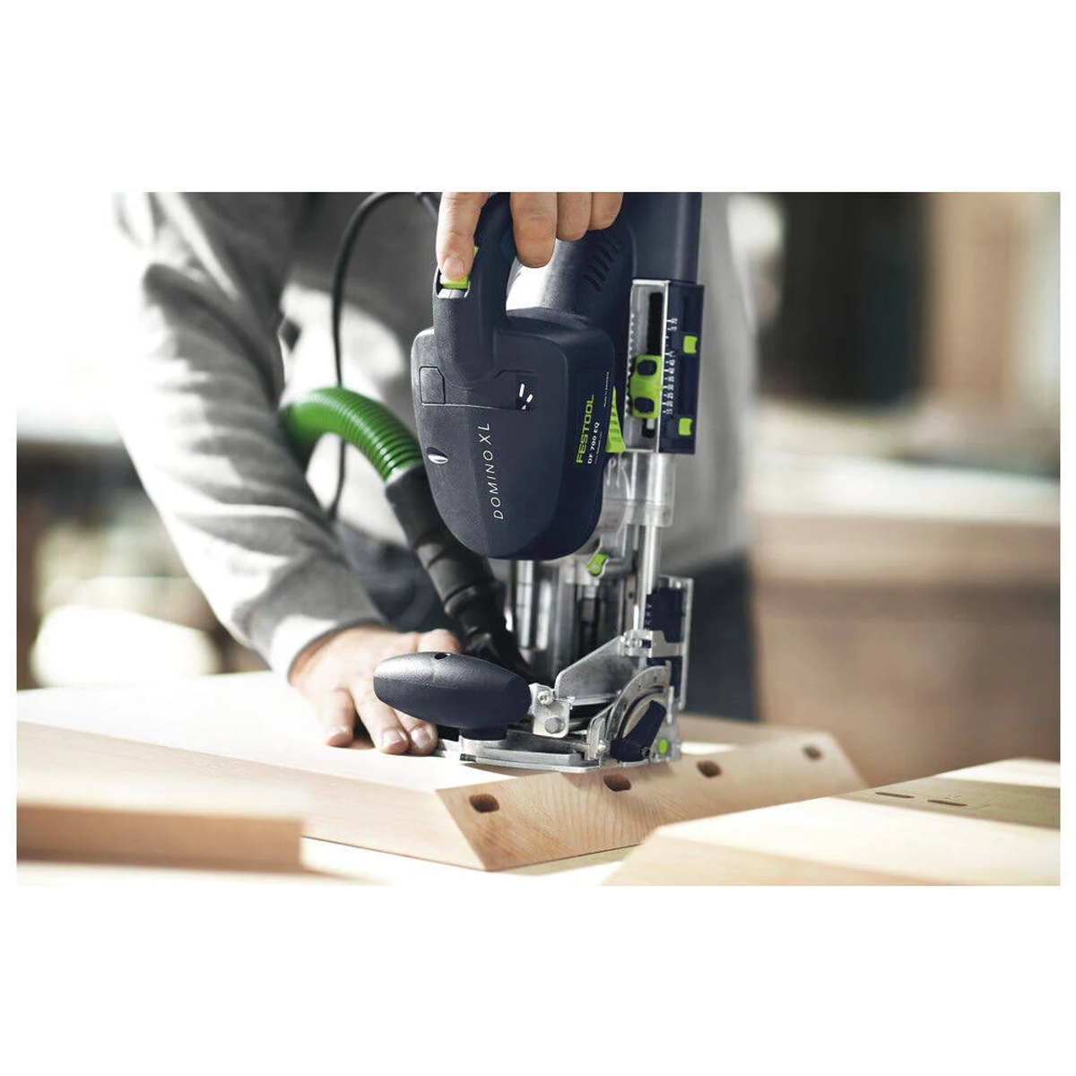 Una persona che utilizza una giuntatrice Festool Domino XL su legno, con un tubo verde collegato, su una superficie bianca.