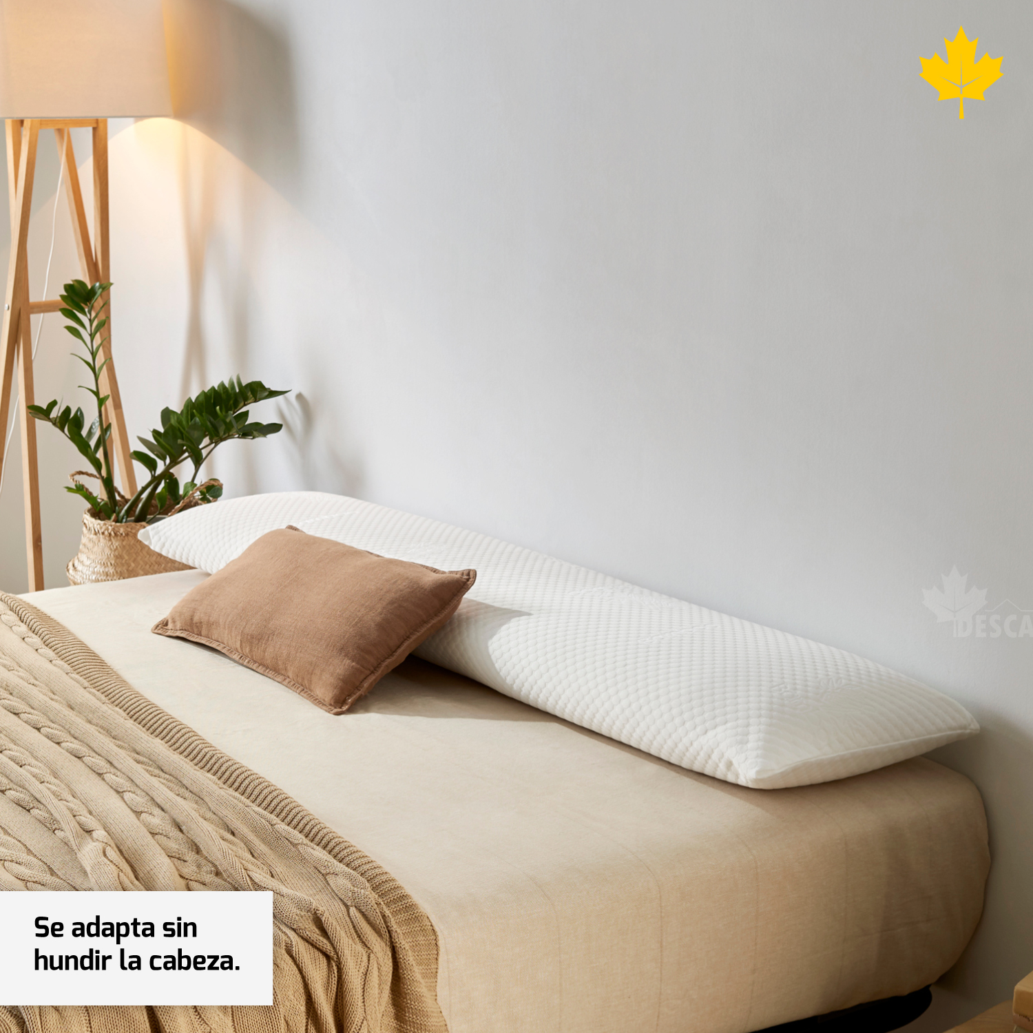 Una almohada larga blanca y una almohada marrón sobre una cama beige, cerca de una lámpara de pie.