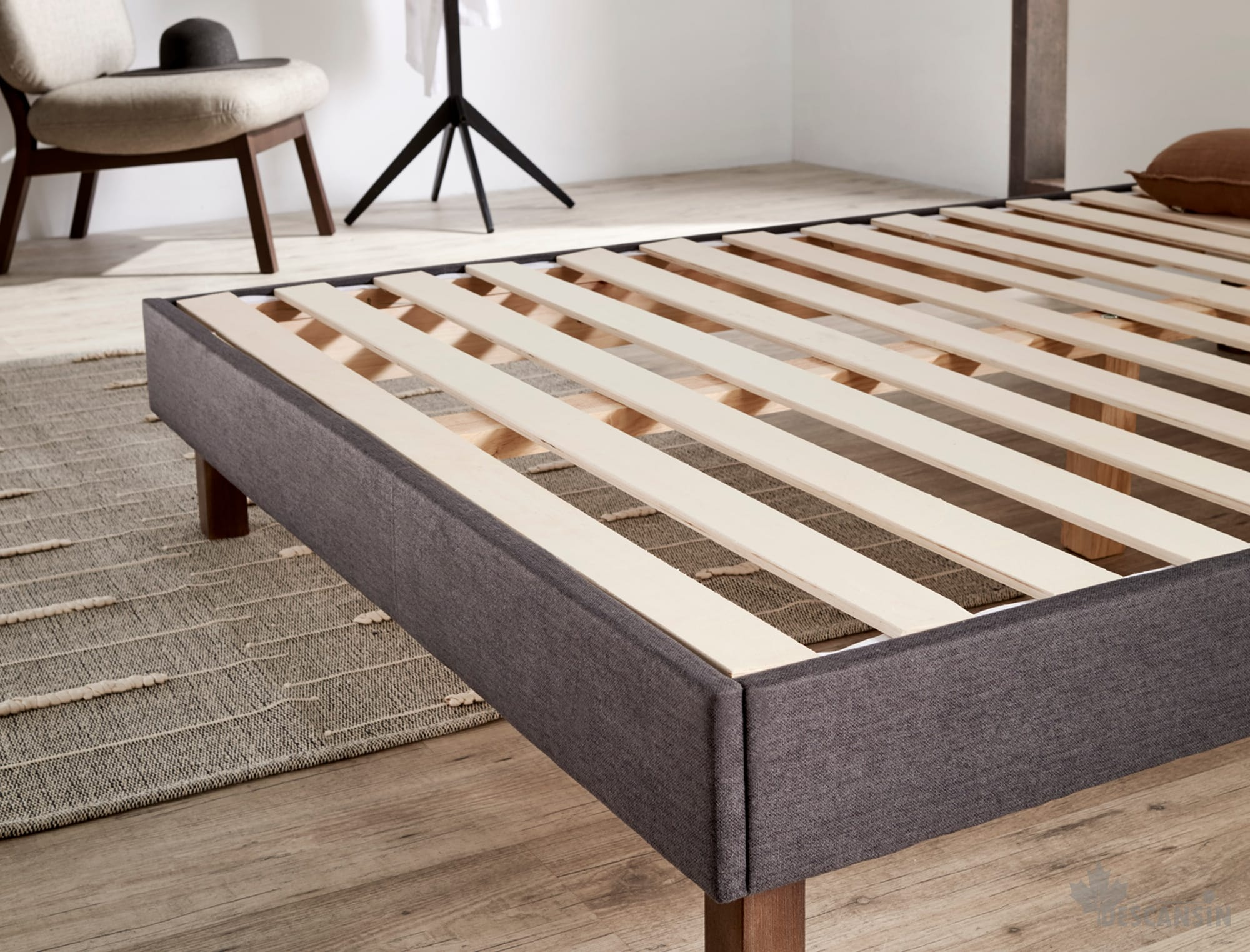 Un marco de cama con listones de madera y tela gris. También se ven una silla y un sombrero.