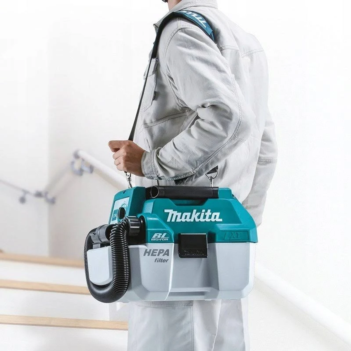 A person carries a teal and gray Makita vacuum cleaner in a white room, walking up stairs.