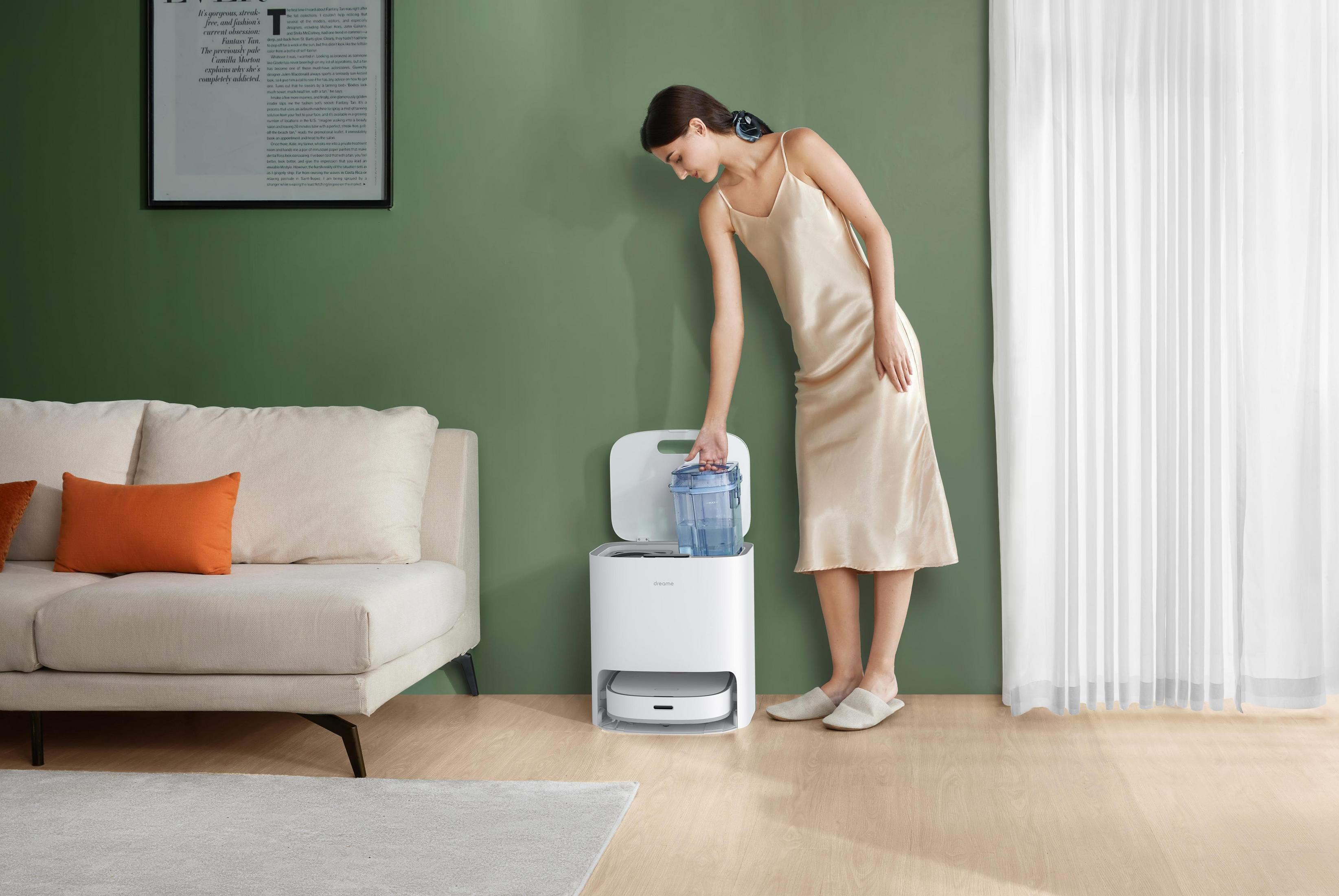 Frau füllt den Wassertank eines weißen Roboterstaubsaugers. Im Hintergrund Sofa, Wandkunst und grüne Wand.