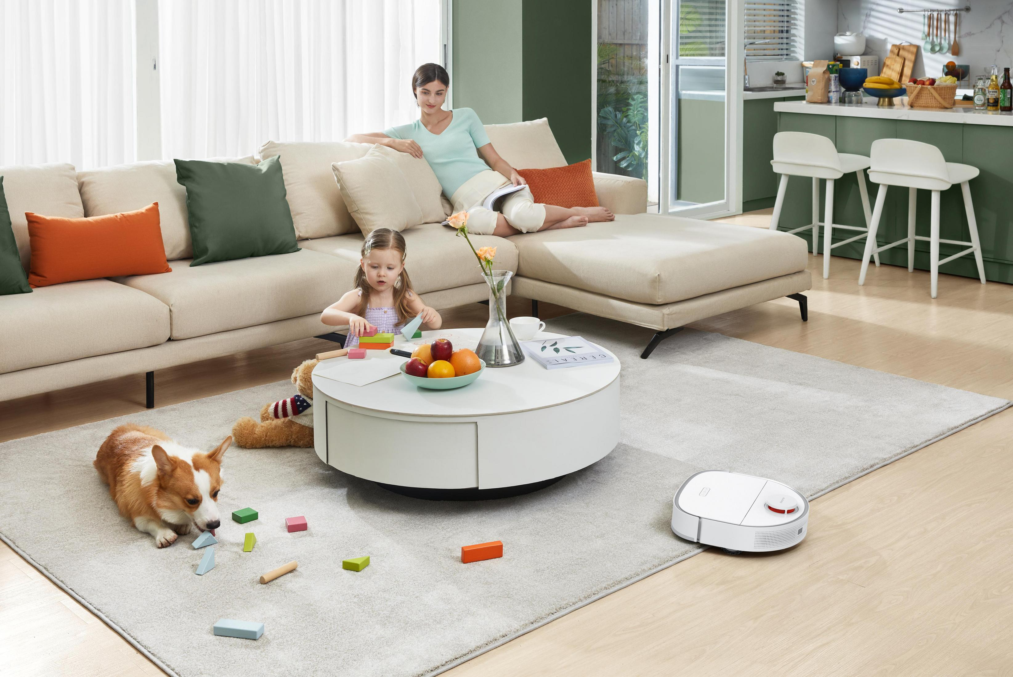 Eine Frau und ein Kind sind in einem Wohnzimmer. Ein Hund und ein Roboterstaubsauger sind vorhanden.