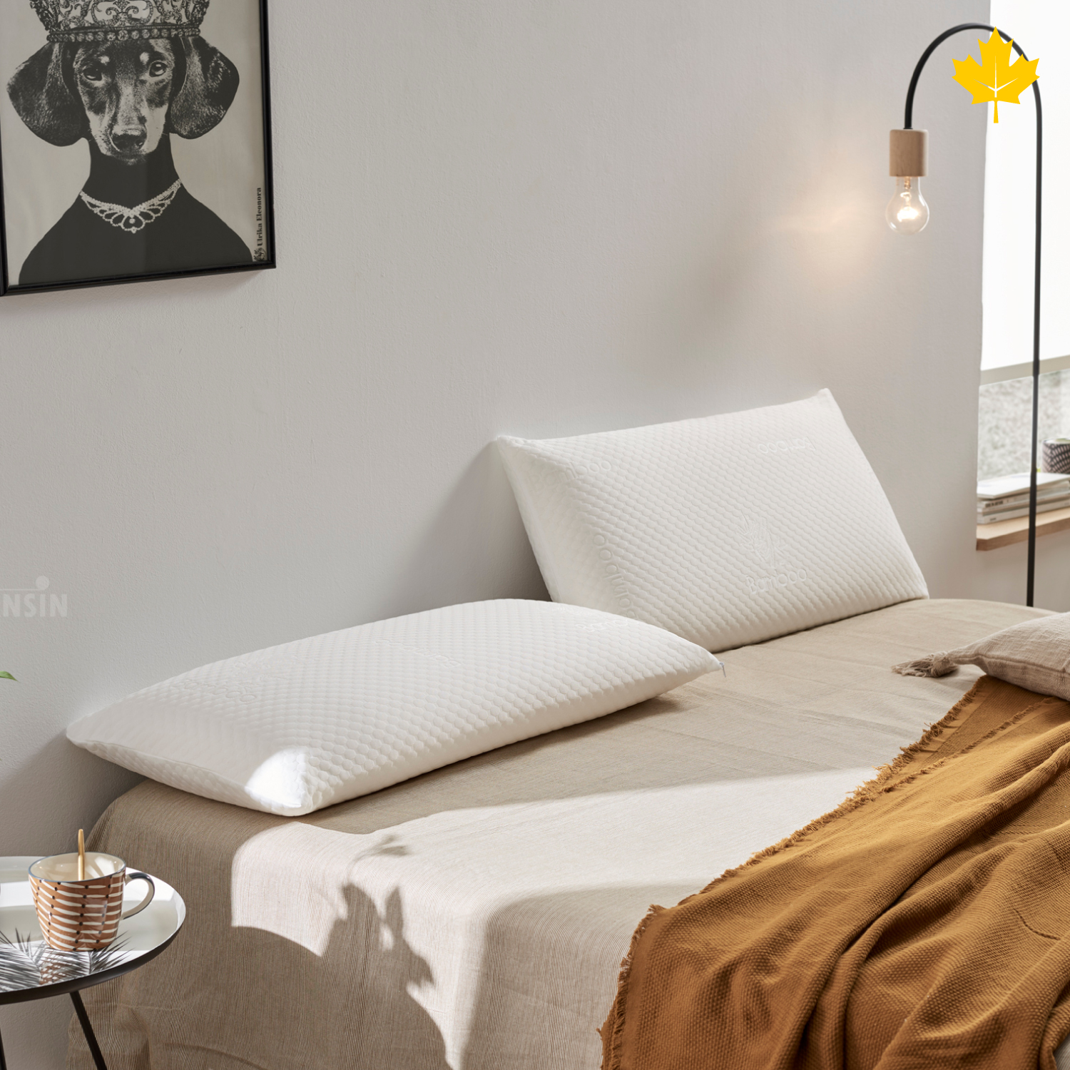 Dos almohadas blancas sobre una cama con sábanas beige y una manta amarilla. Visible una mesita y un cuadro.