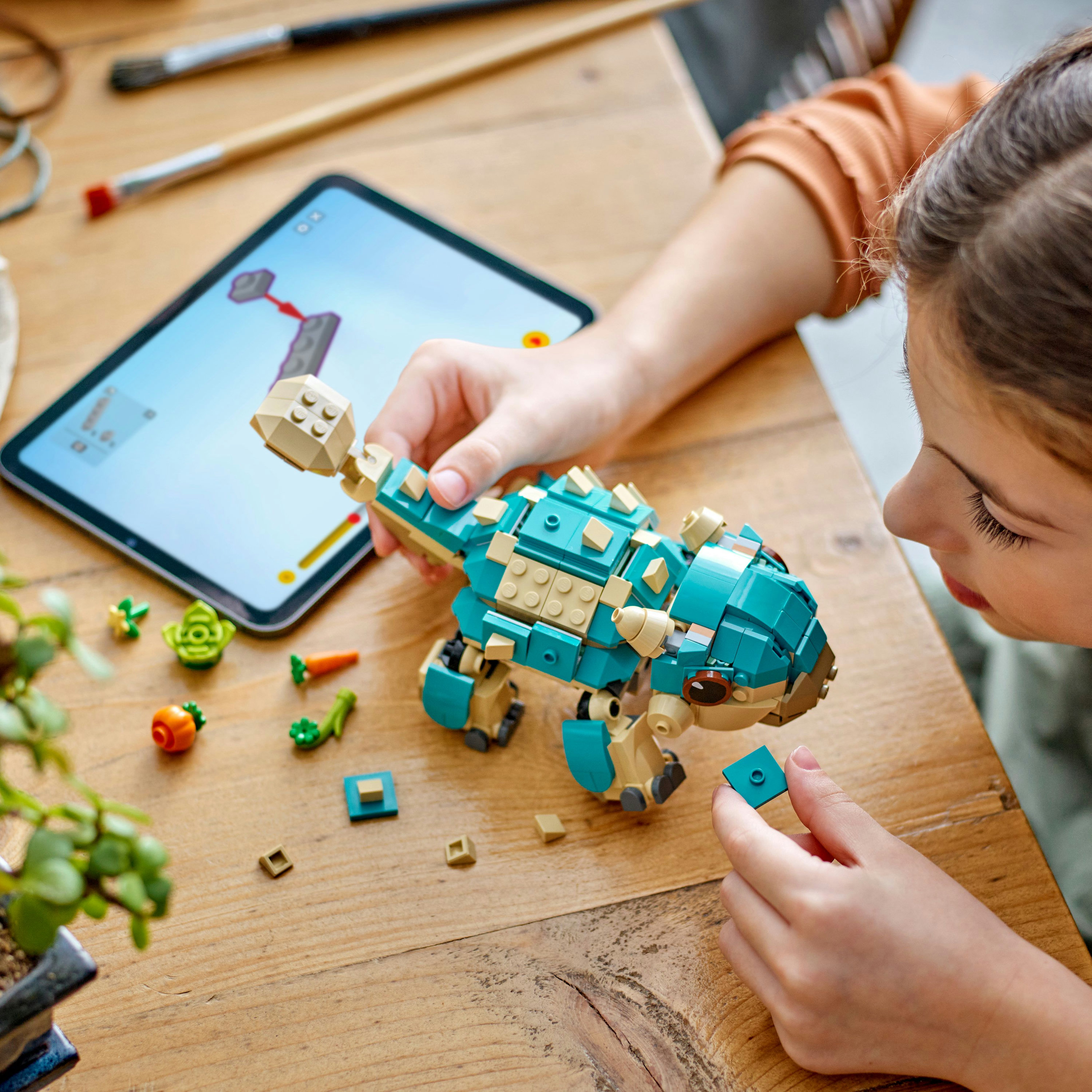 Un niño construye un dinosaurio Lego. Una tableta está abierta. Piezas de Lego están esparcidas.