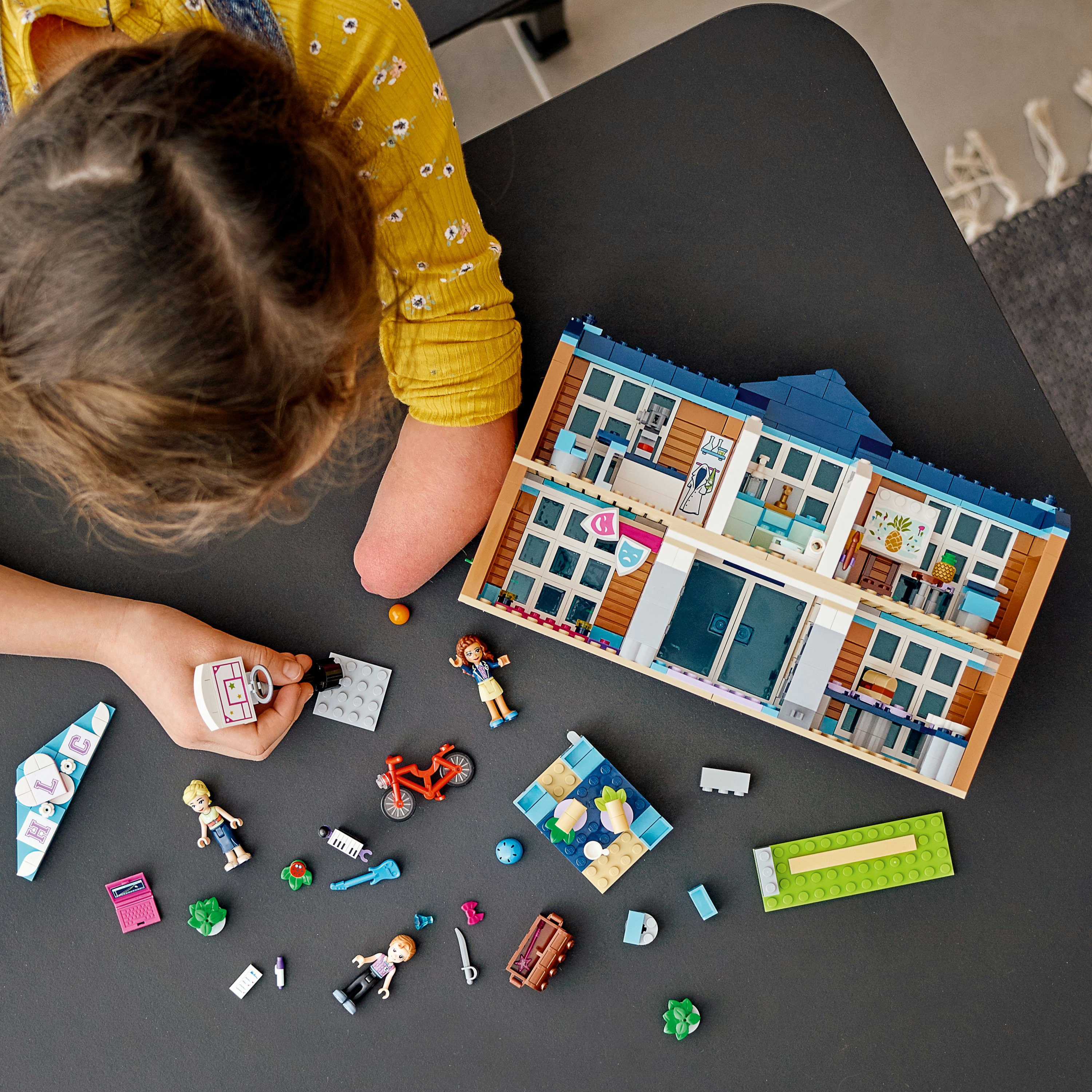 Ein Mädchen spielt mit einem Lego-Set. Das Set ähnelt einer Schule. Es gibt kleine Lego-Figuren.
