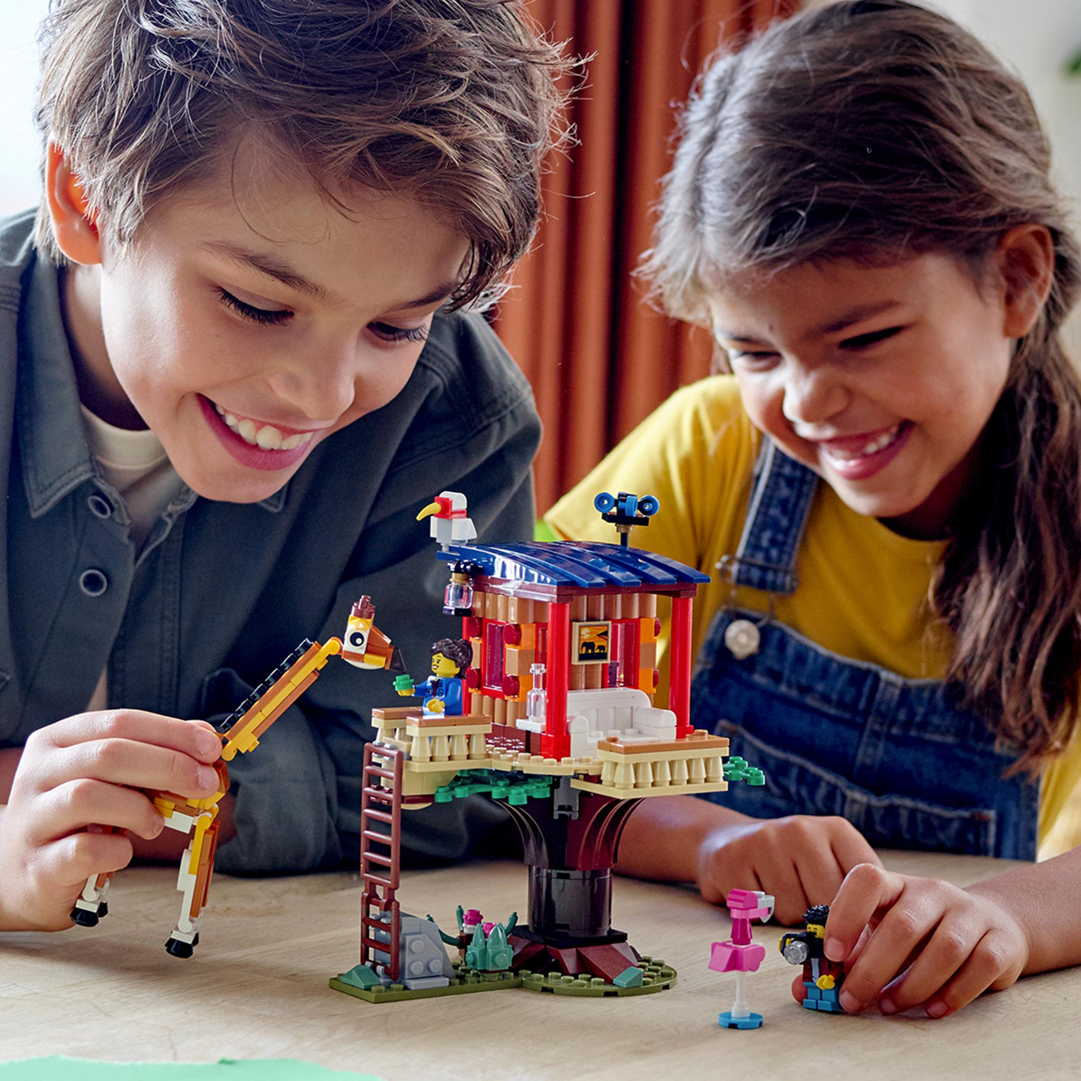 Due bambini giocano con un set LEGO. Una casa sull'albero con una giraffa e un fenicottero è al centro.